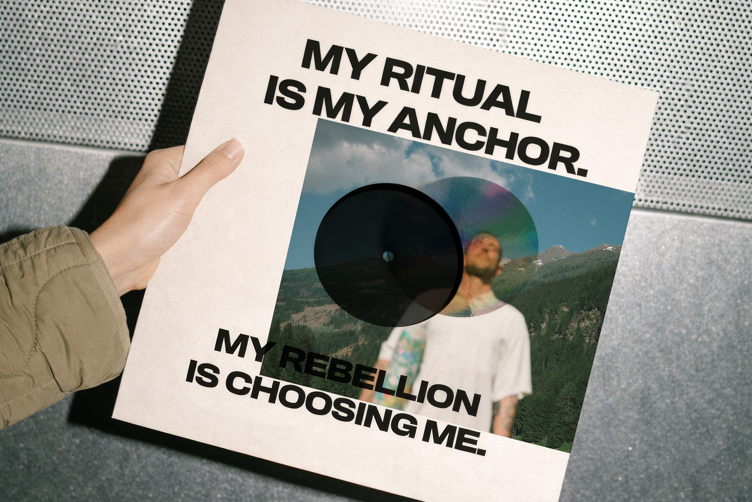 A person holding a sign with a photo of a landscape, a vinyl record, and text that reads "My ritual is my anchor. My rebellion is choosing me." on a textured surface.