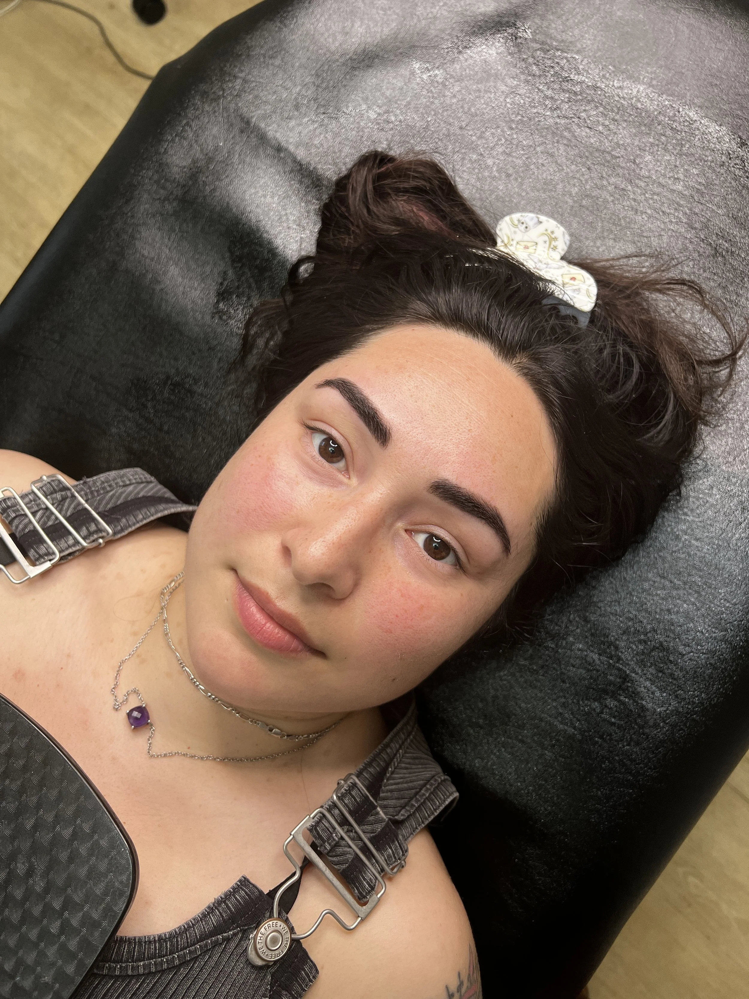 A young woman lying on a black leather table, with her head resting on a black cushioned headrest, looking at the camera.