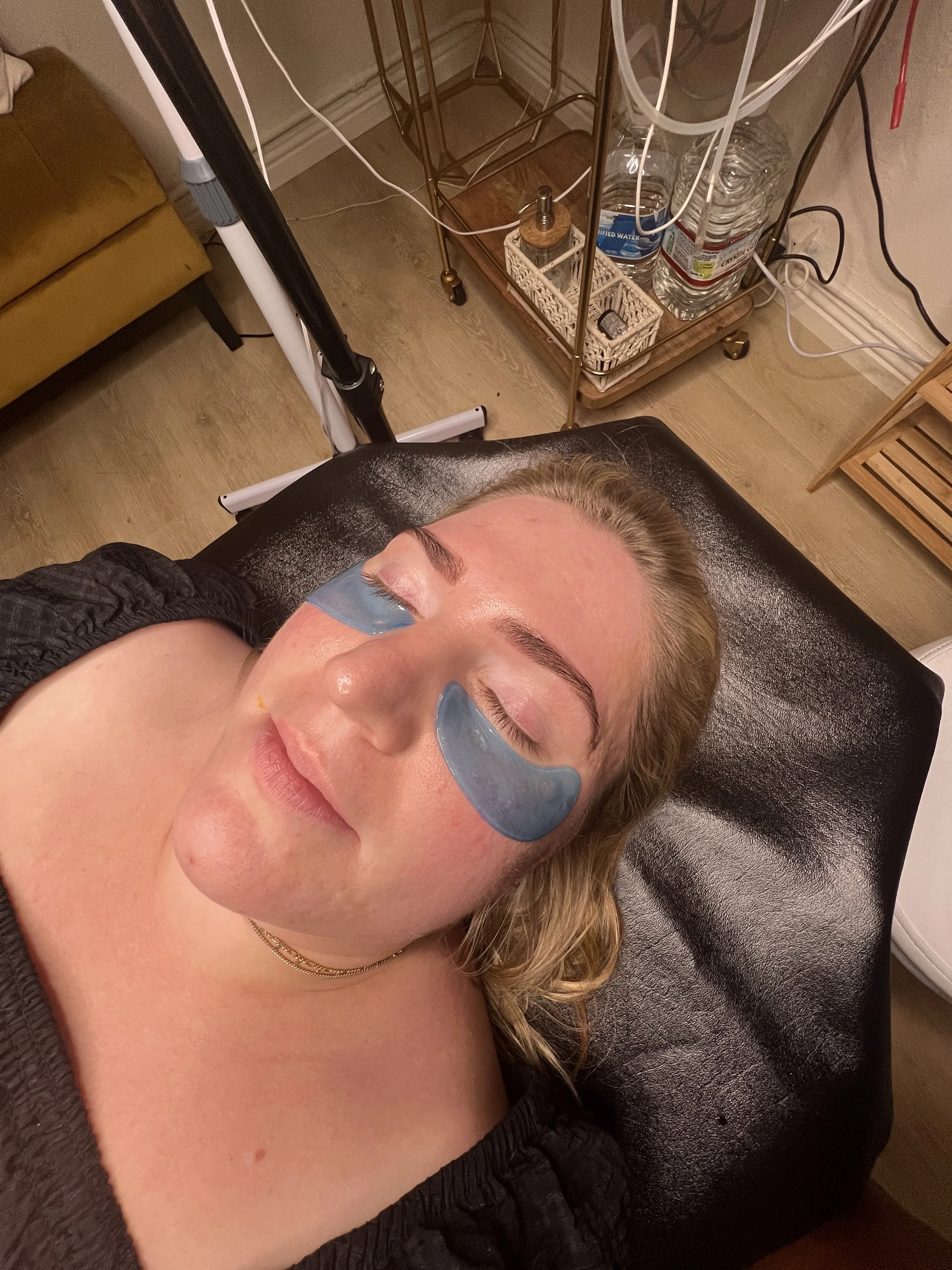 A woman with eye patches lying on a treatment table, with a relaxed expression, in a spa or skincare clinic setting.
