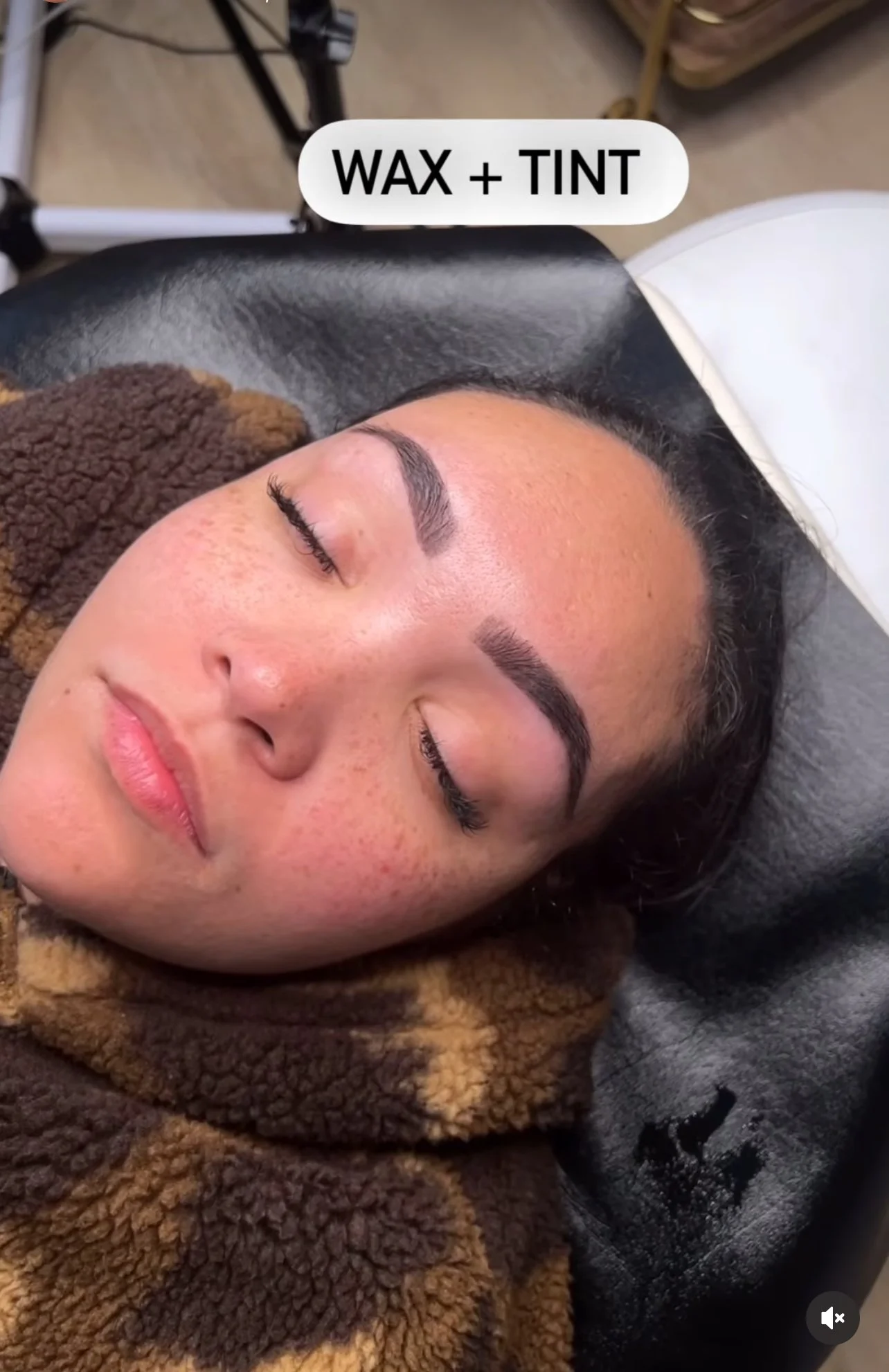 A woman lying on a salon chair with her eyes closed, receiving a waxing and tinting procedure on her eyebrows, with a caption indicating 'WAX + TINT'.