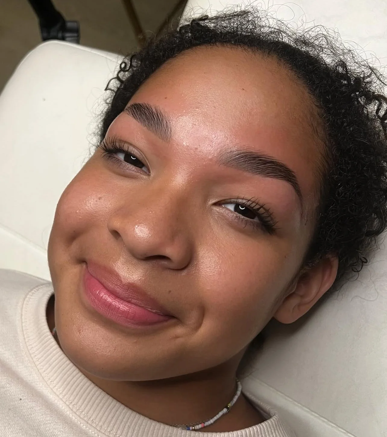 Close-up of a young woman with curly hair lying on a white medical chair, smiling at the camera.