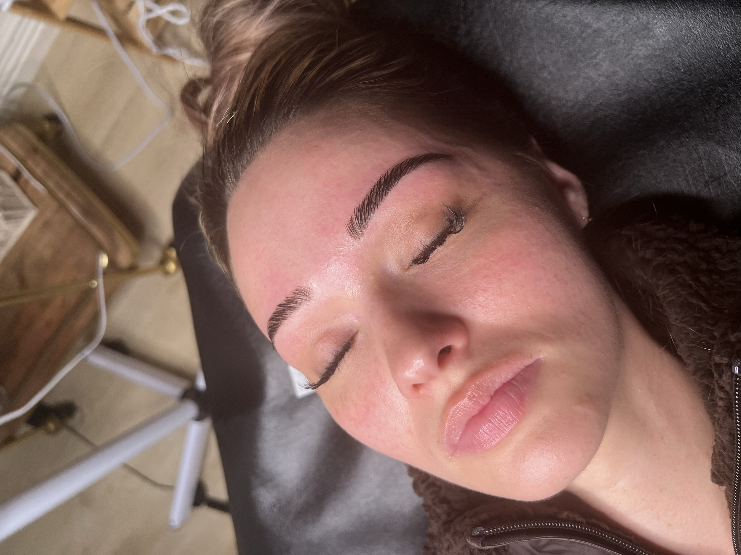 A woman with makeup and manicured eyebrows lying with her eyes closed, possibly at a salon or beauty clinic.