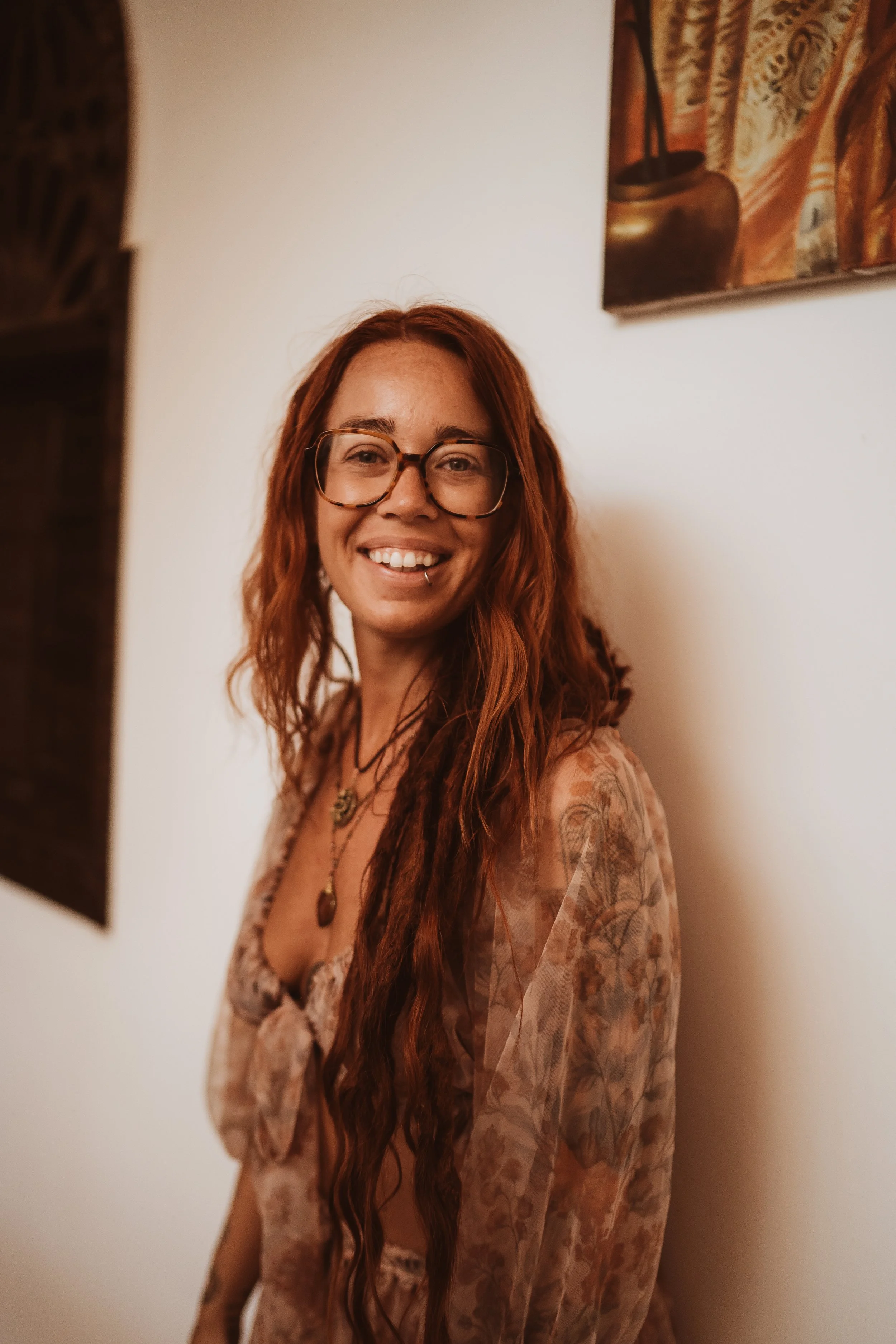 A young creative woman with long, wavy red hair, glasses, and a piercing smiling at the camera, standing against a white wall with artwork.