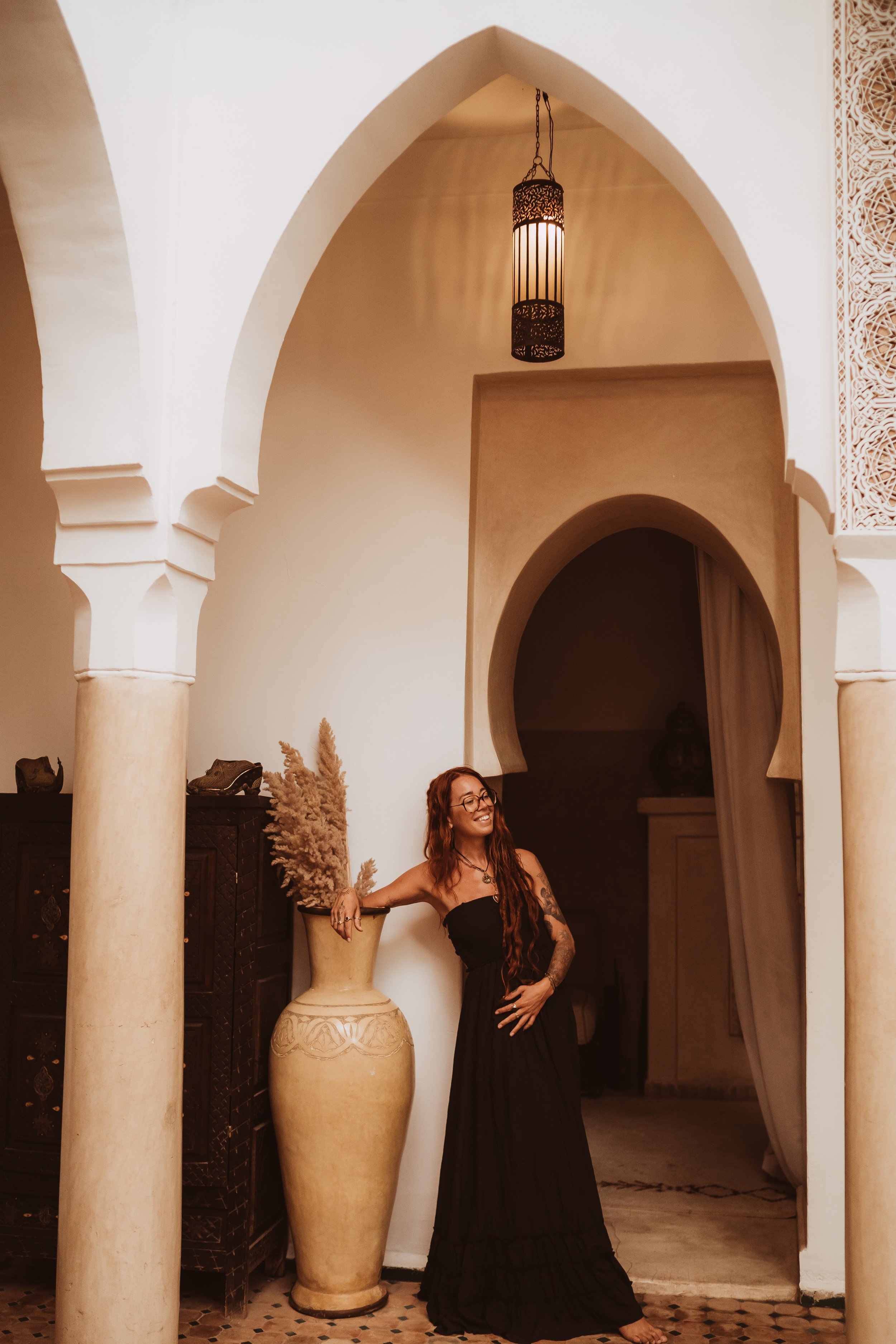 Woman with long red hair in a black strapless dress standing next to a large vase with pampas grass, inside an ornately decorated room with arches and a hanging lantern.