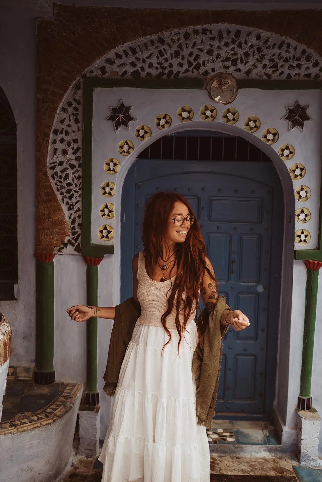 A woman with long red hair, glasses, and tattoos standing in front of an ornate door with decorative tiles and arches, smiling and looking to her right.
