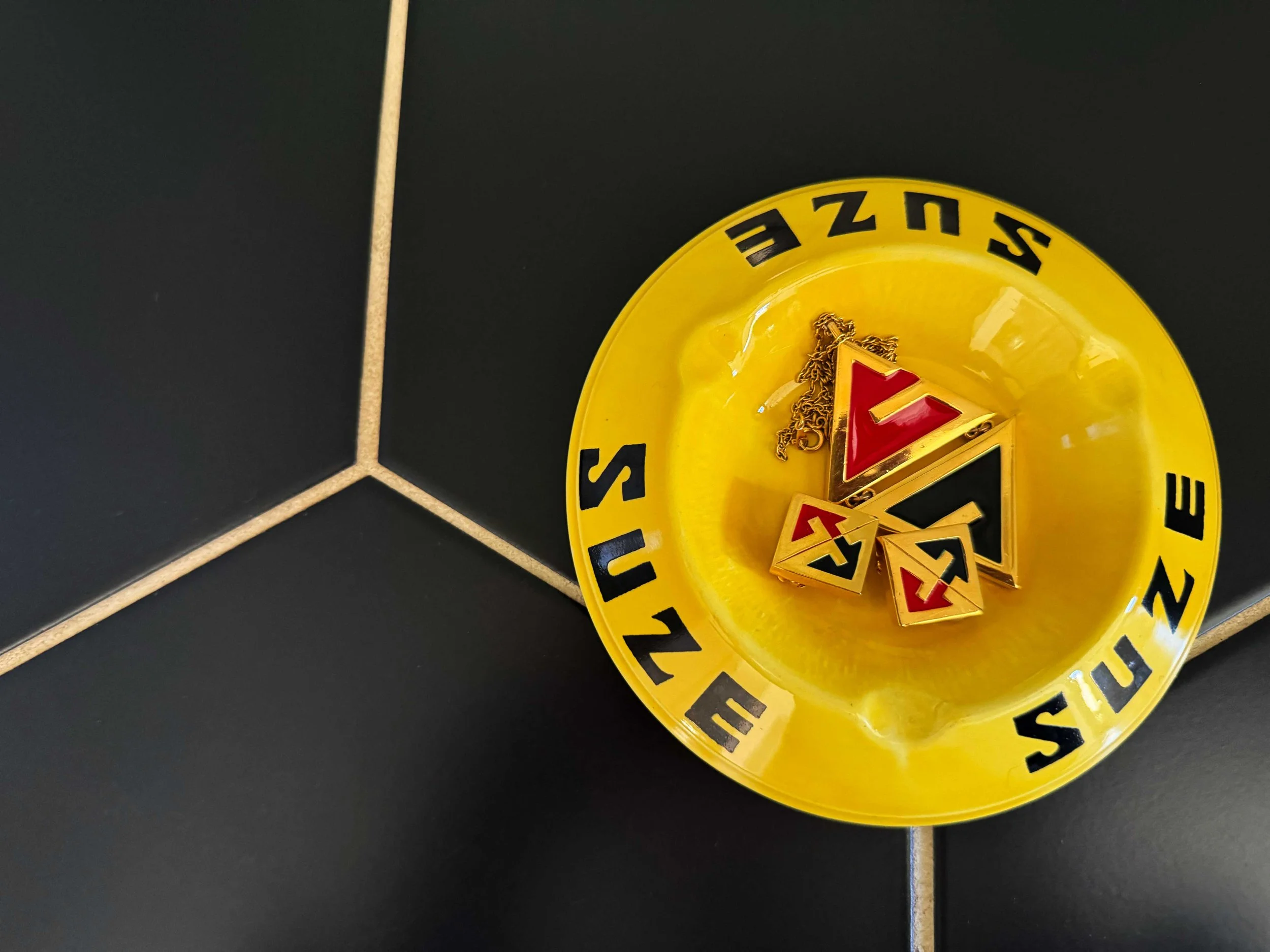 Yellow ashtray with the words "Suzee" and "Eauze" printed in black, containing jewelry including a triangular pendant and other small pieces.