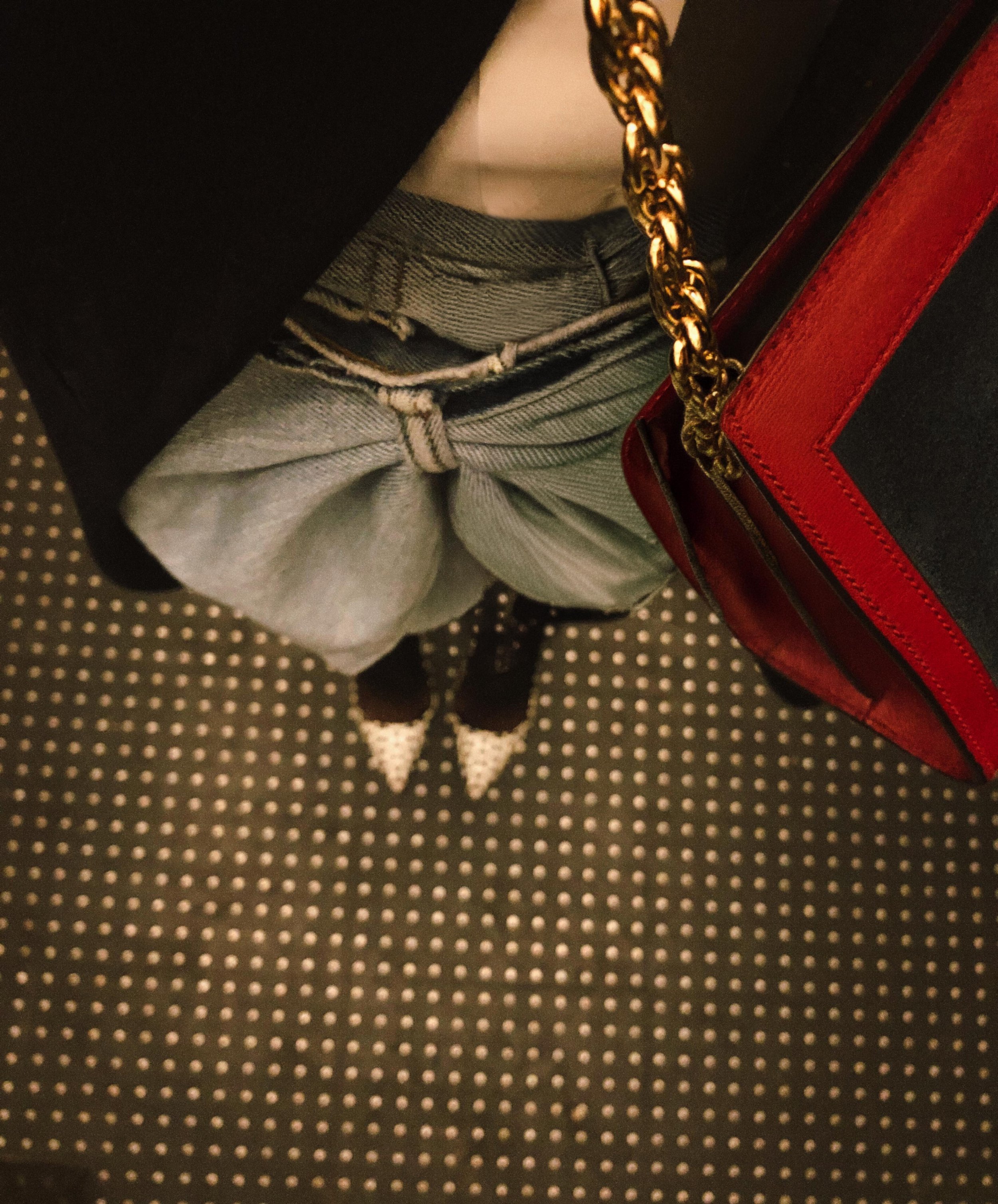 Person wearing gray sweatpants with frayed waistband, black shoes with white capped toes, holding a red handbag with a gold chain strap, seen from an overhead perspective standing on a dotted carpet.