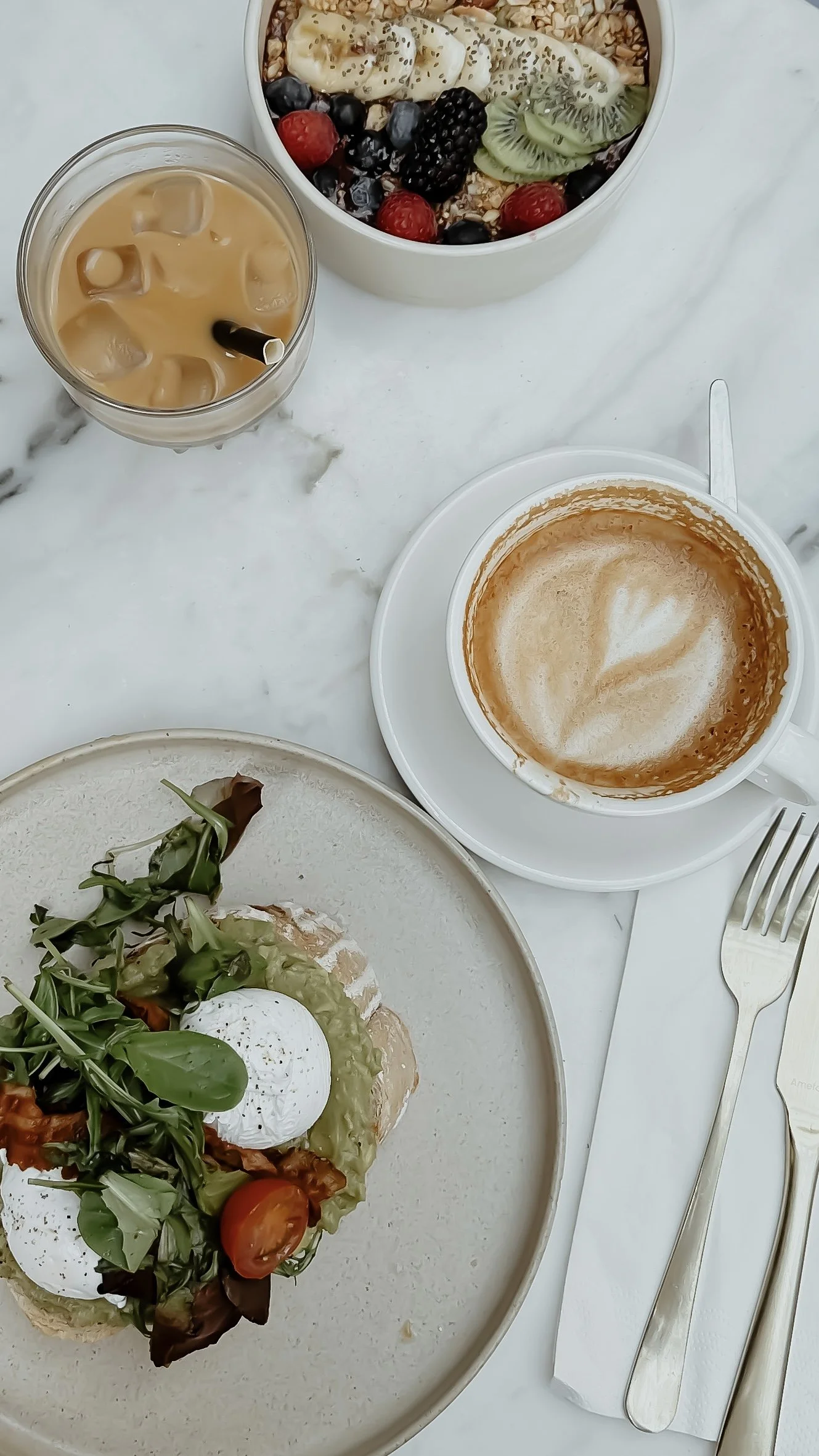 En tallrik med ägg Benedict med grönsaker och matolja, en kopp med cappuccino, ett glas med iced latte, en skål med yoghurt eller smoothie toppad med färska bär och kiwi, samt en gaffel och kniv.