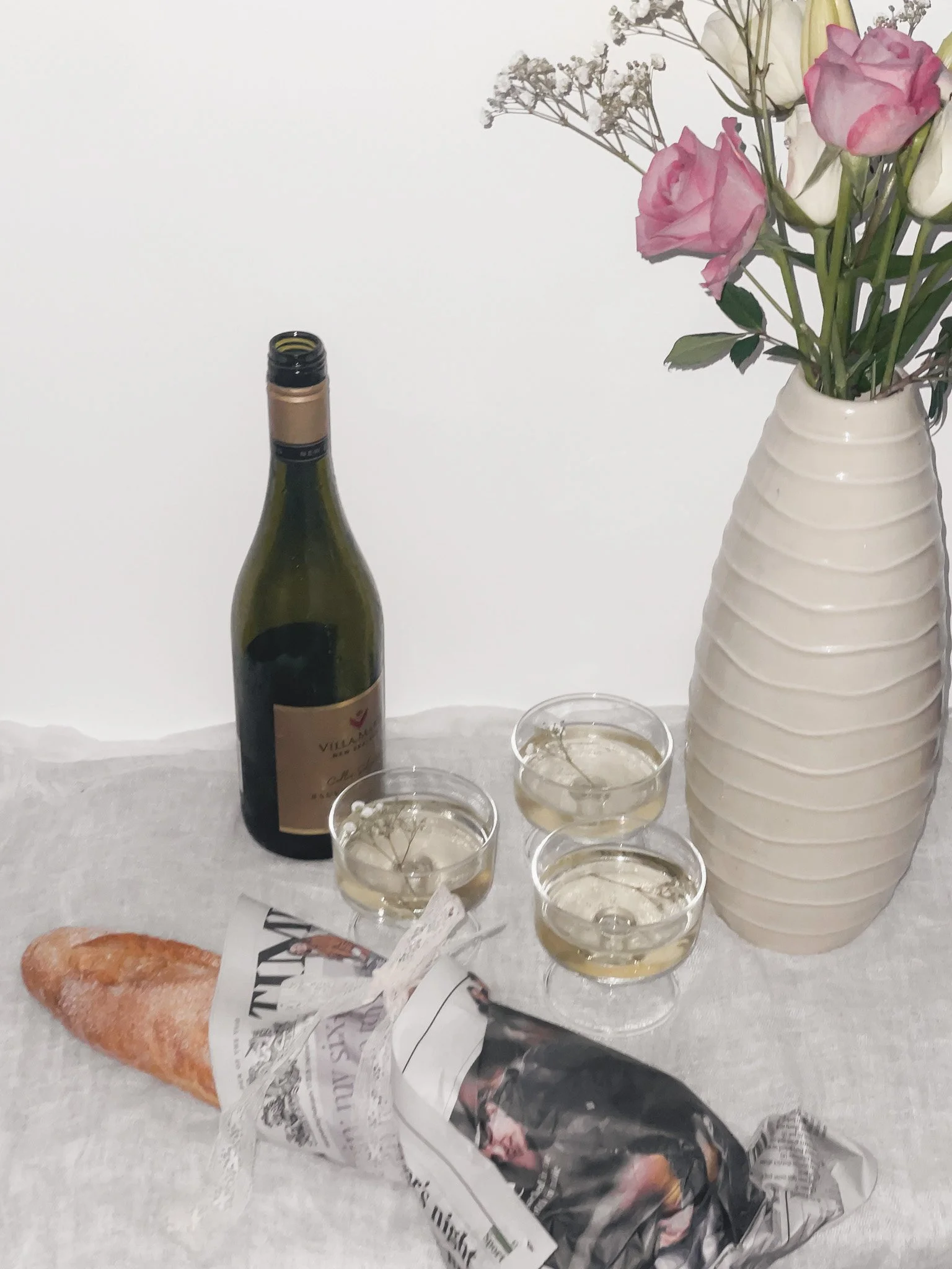 En tom vinflaska, tre vinglas med vitt vin, en växt i en vit kakel, ett bakverk i papper, och en vas med rosa och vita blommor.