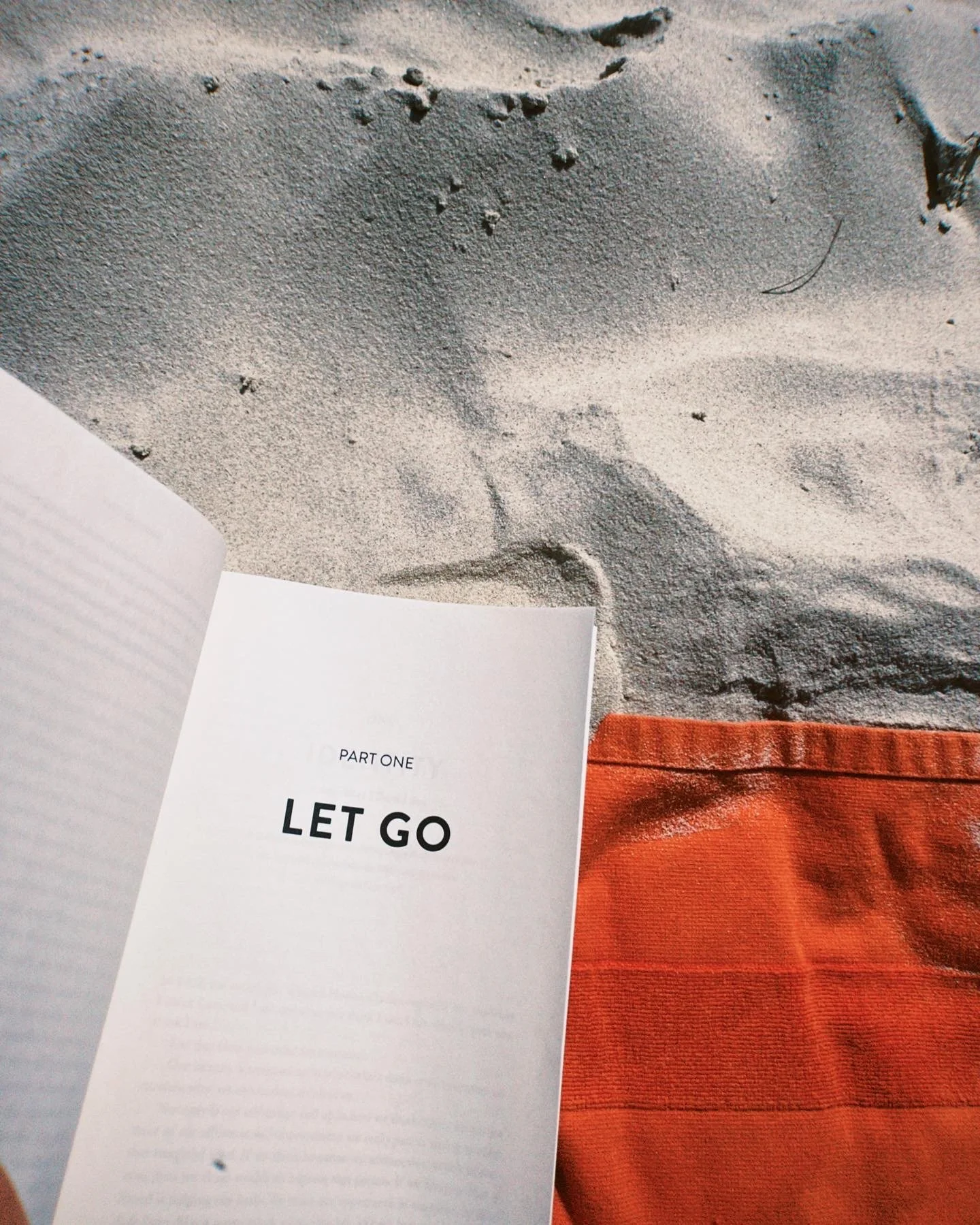 Bok med titeln 'Part One: Let Go' på sand vid stranden, med en orange tygpåse i närheten.
