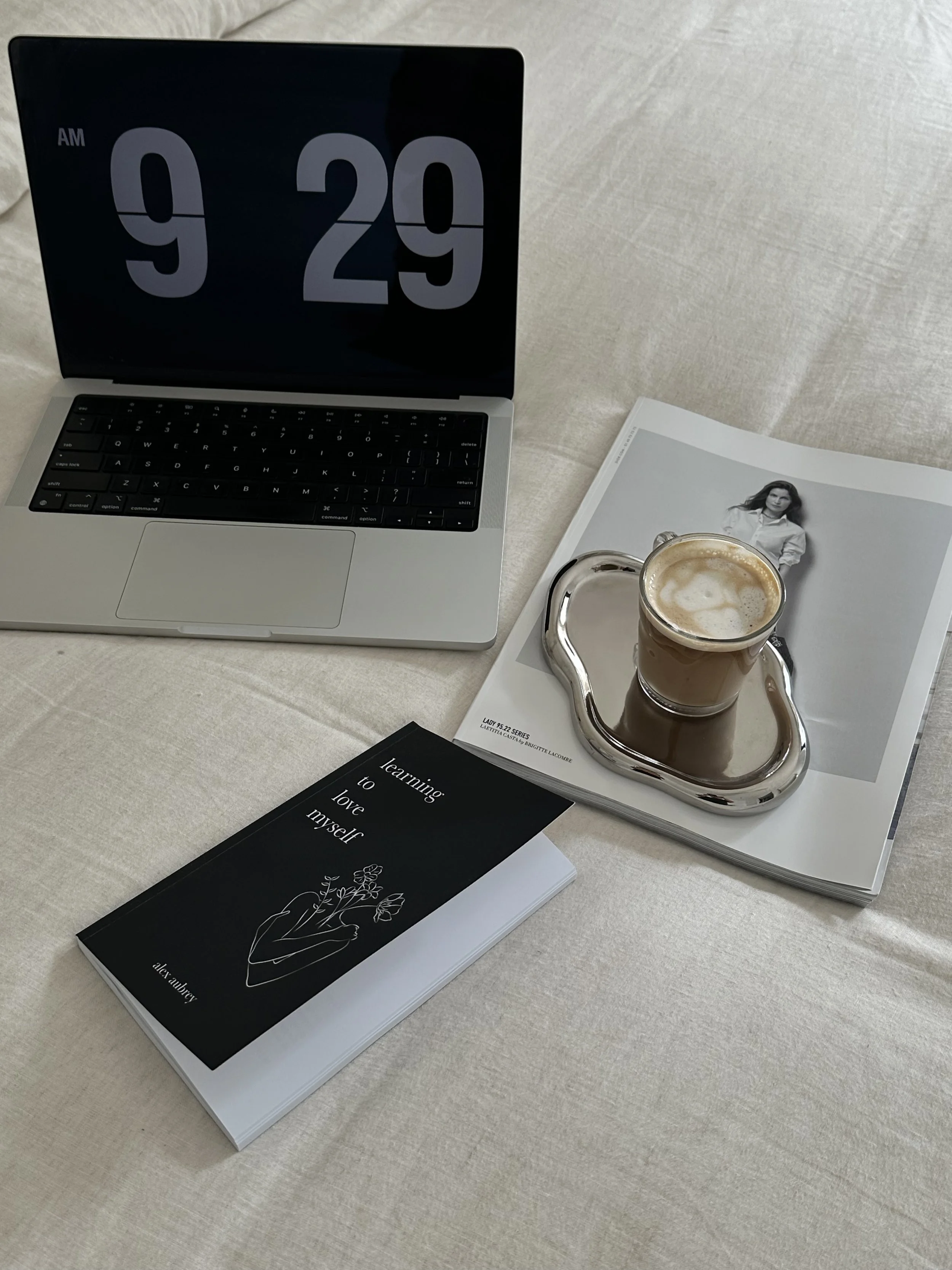 En öppen bärbar dator med stor digital klocka som visar 9:29. På bänken finns en kopp kaffe på ett silverfat, en svart bok med titeln "learning to love myself" av alex armey, och ett magasin med en svartvitt bild av en kvinna.
