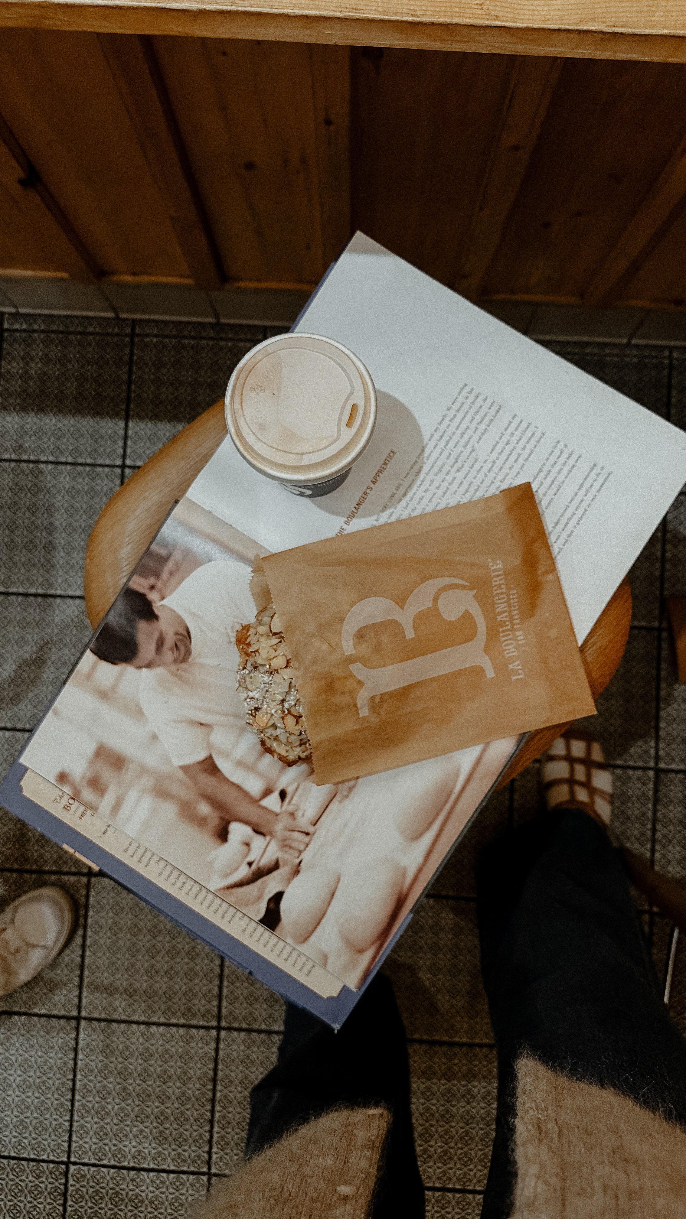 En kopp kaffe, en muffins i papperspåse med texten 'L A DOULEUR?' och en bok eller broschyr på ett runt bord, taget från ovan.