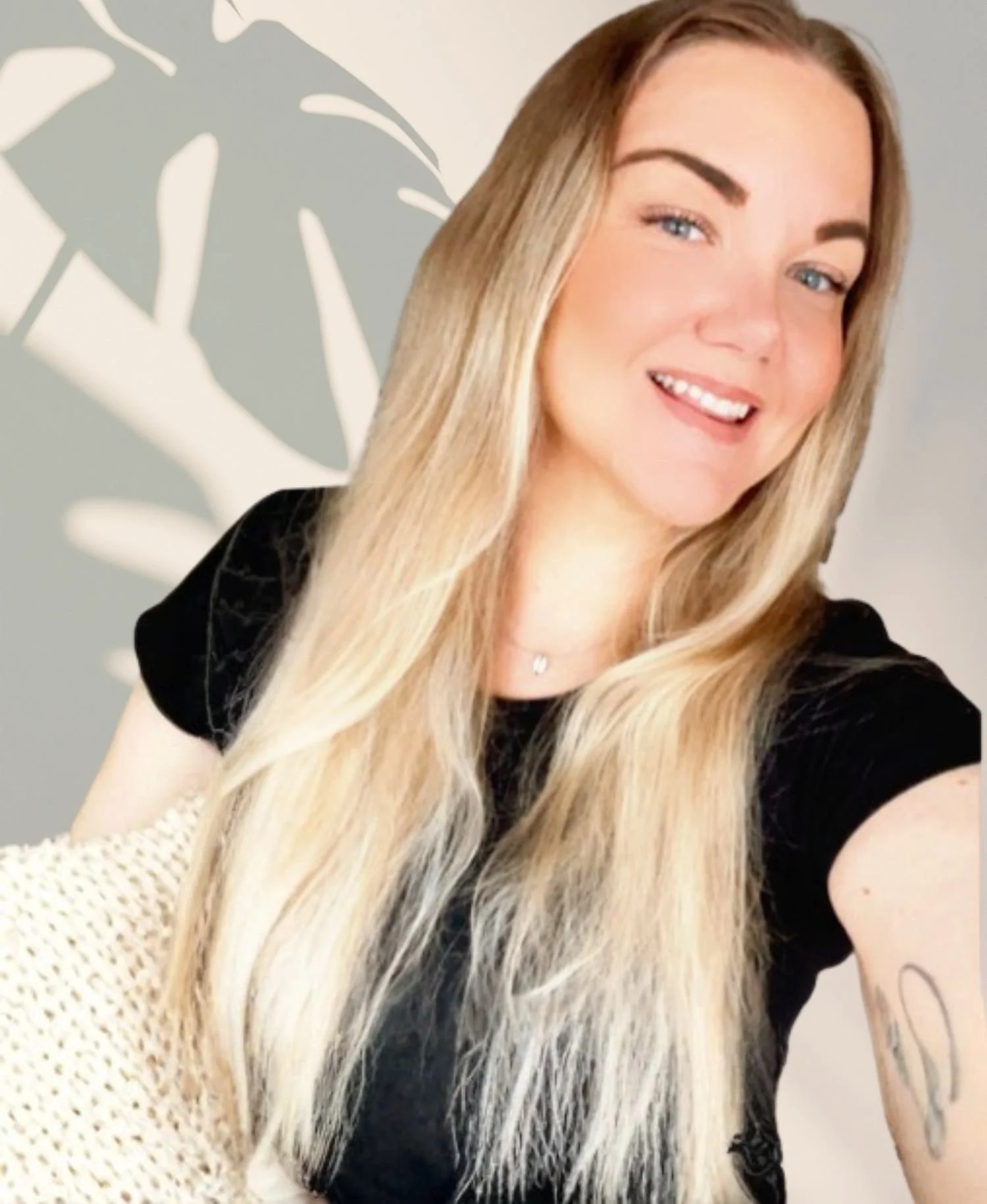 En ung kvinna med långt blont hår, svart t-shirt, som ler mot kameran. Bakgrund av en vägg med ett stort, grått bladmönster.