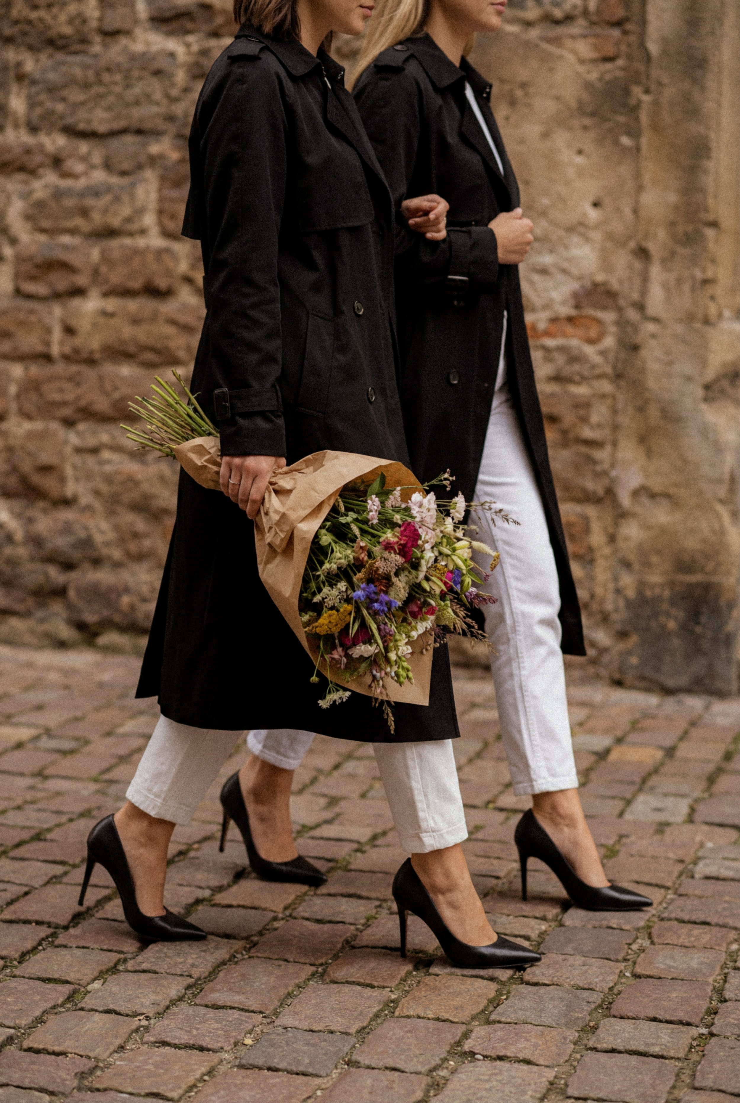 Två kvinnor går på en gatusträcka intill en gammal tegelvägg, den ena bär en blomsterbukett och båda är klädda i svarta kappor och svarta högklackade skor.