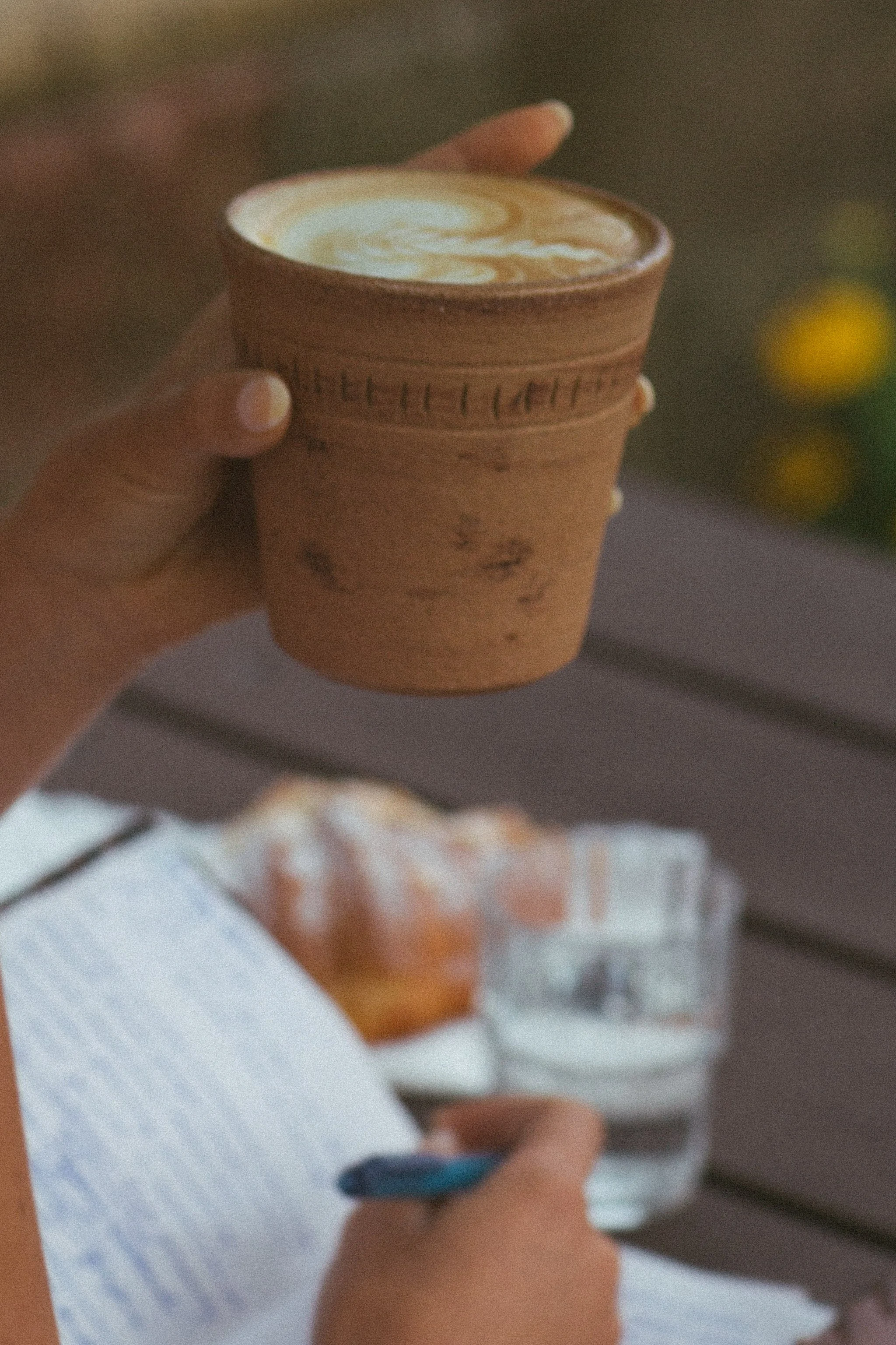 En person håller ett kopparlock med en varm dryck, eventuellt kaffe eller latte, framför en bänk med en brödlimpa med smör och en kopp kaffe och en hand som skriver i en anteckningsbok.