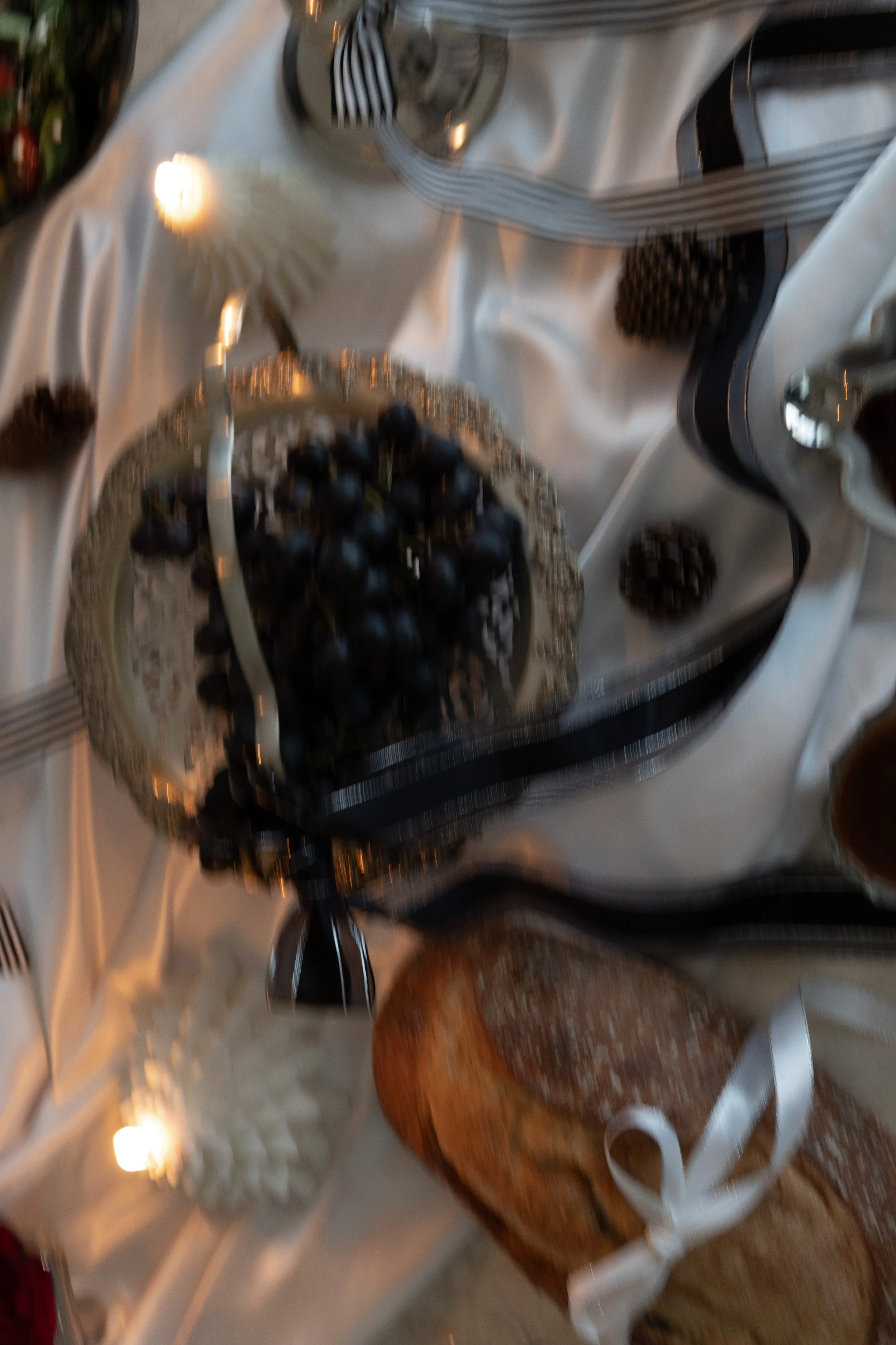 Blurrigt foto av ett middagsbord med vinbär i en glasfat, ett bröd med vit band, ett ljus, och andra tillbehör på en vit duk.