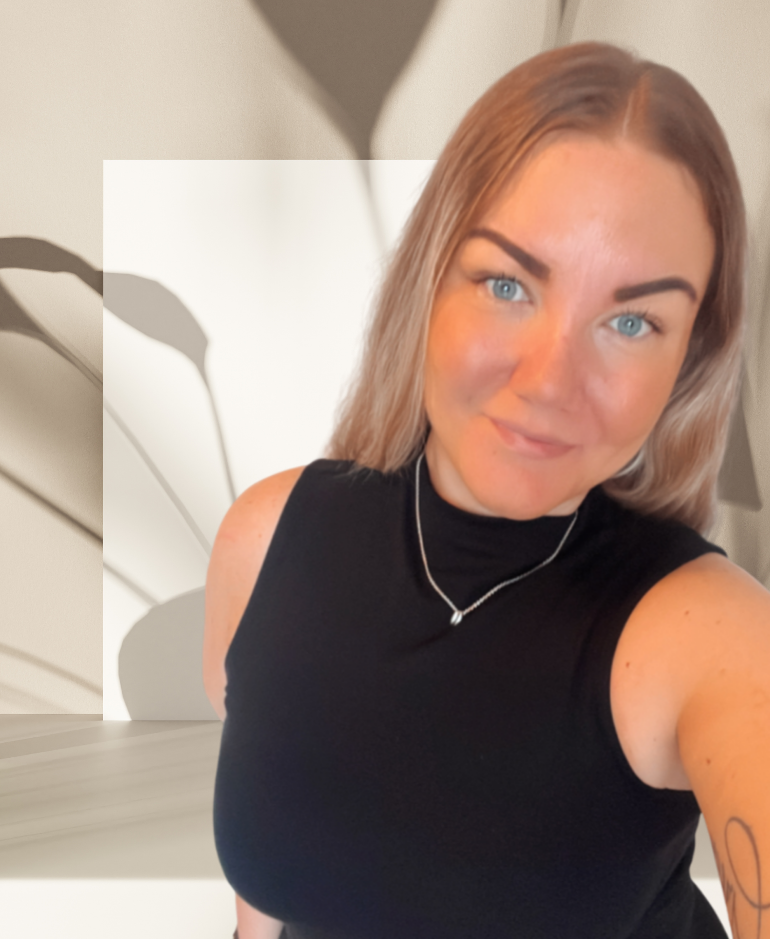 Ung kvinna med ljusbrunt hår, blå ögon, svart ärmlös topp, silverhalsband och tatuering på armen, som tar en selfie framför en vägg med skuggor.