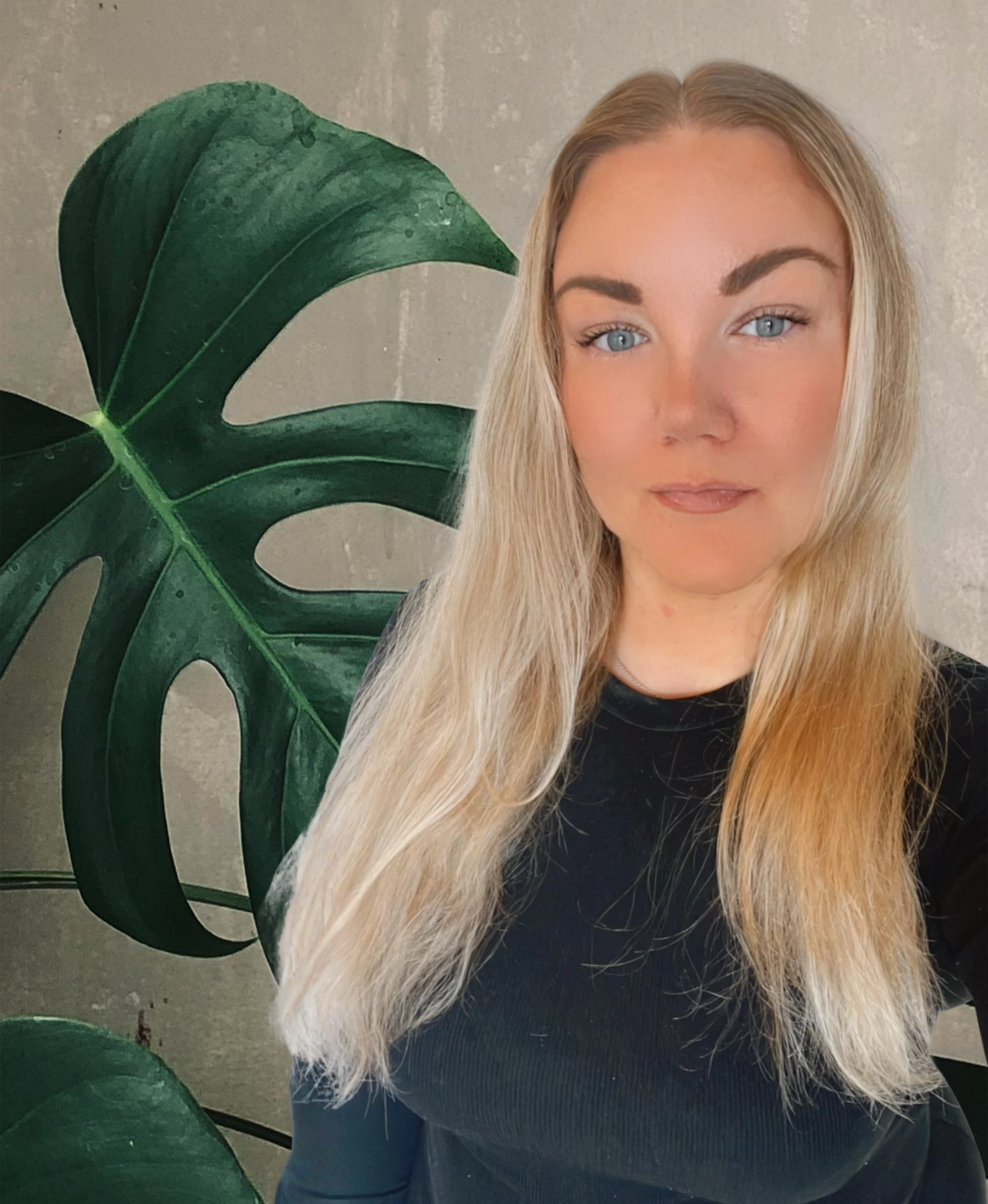 En ung kvinna med långt blont hår och blå ögon står framför en växt med stora gröna blad. Hon bär en svart tröja och tittar mot kameran.