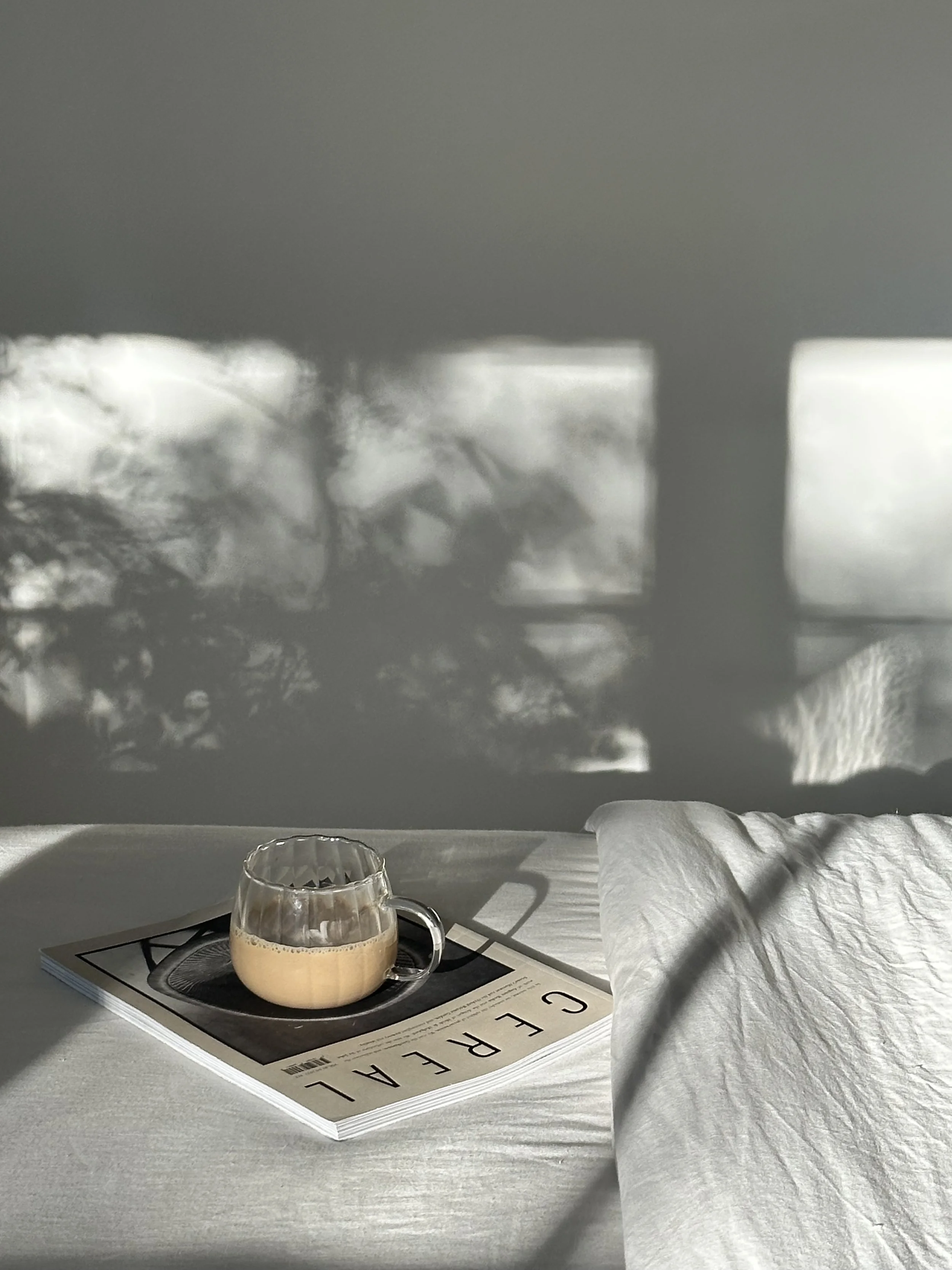 Ett glas cappuccino på en tidning med en vit säng och ljus som reflekteras i fönstret.