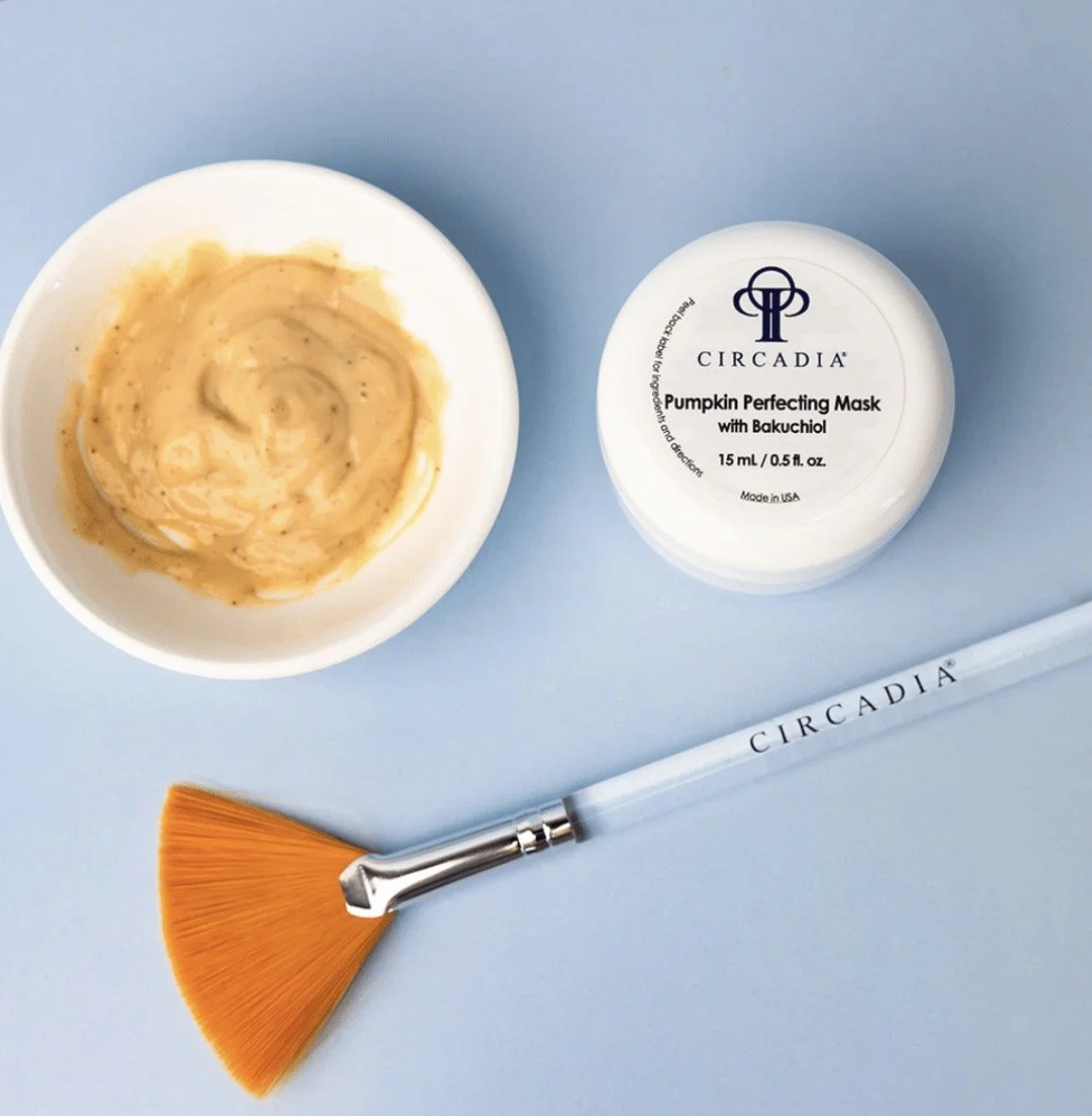 A collection of skincare tools and products on a blue surface, including a white bowl with a yellow-orange facial mask, a jar of Circadia Pumpkin Perfecting Mask, a small fan brush with "CIRCADIA" written on the handle.