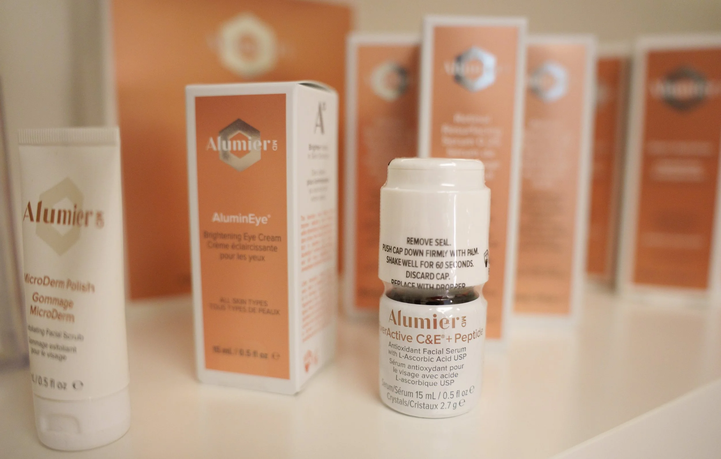 A small bottle of Alumier MD skincare product with a white cap, and several boxes of Alumier skincare products in the background on a white surface.