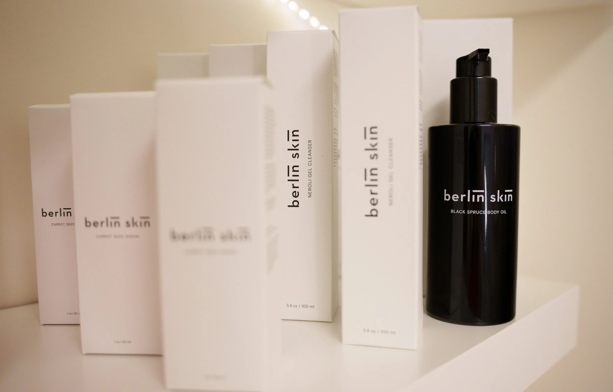 Black bottle of Berlin Skin Black Spruce Body Oil next to white boxes of Berlin Skin skincare products on a white shelf.