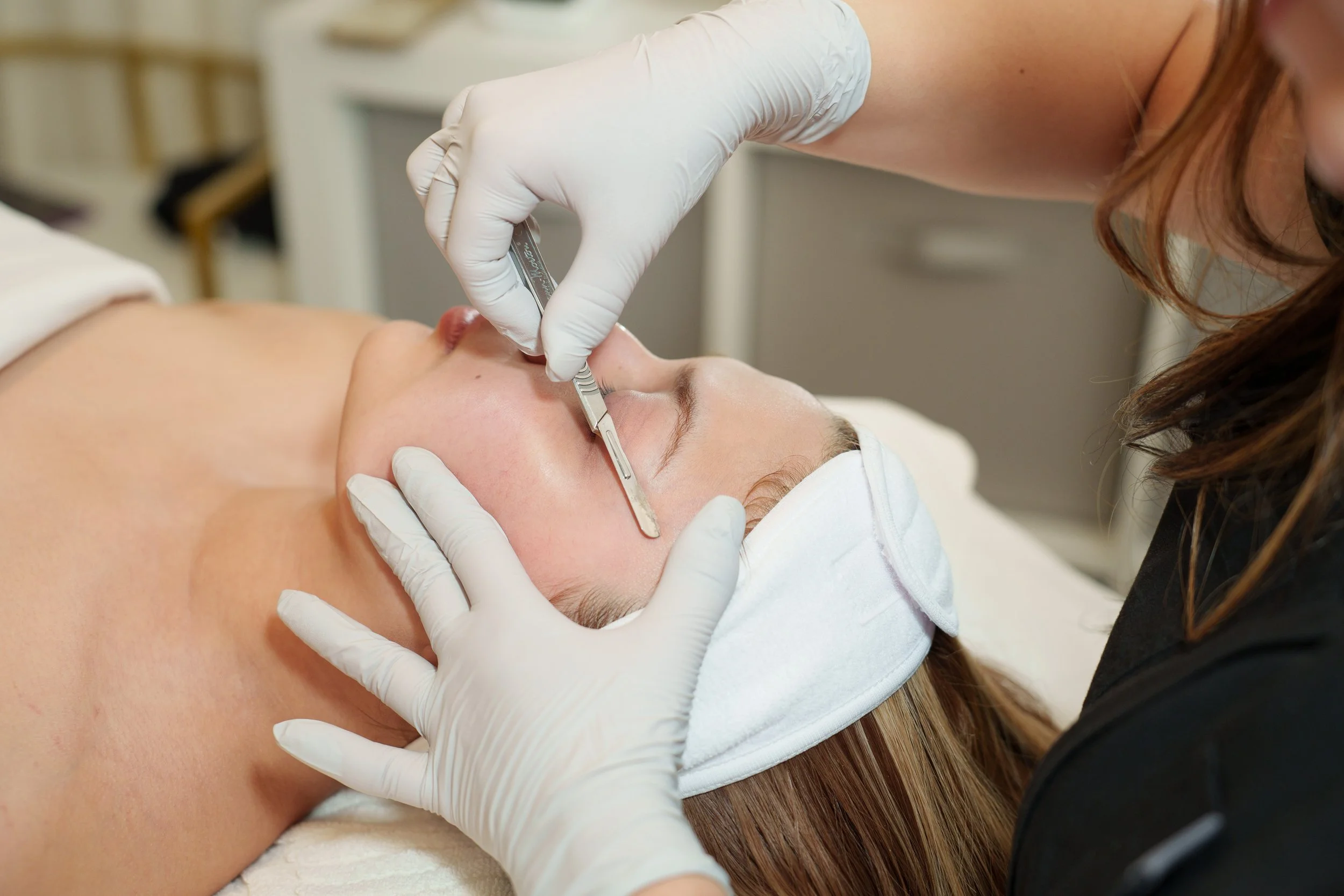A woman is lying peacefully on a treatment bed with her eyes closed, receiving a facial treatment in a spa or clinic setting.