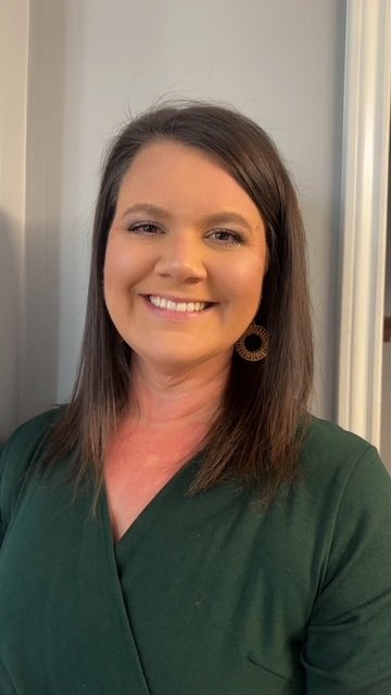 A woman with shoulder-length brown hair smiling, wearing a dark green top and large circular earrings, standing in front of a gray wall and mirror.