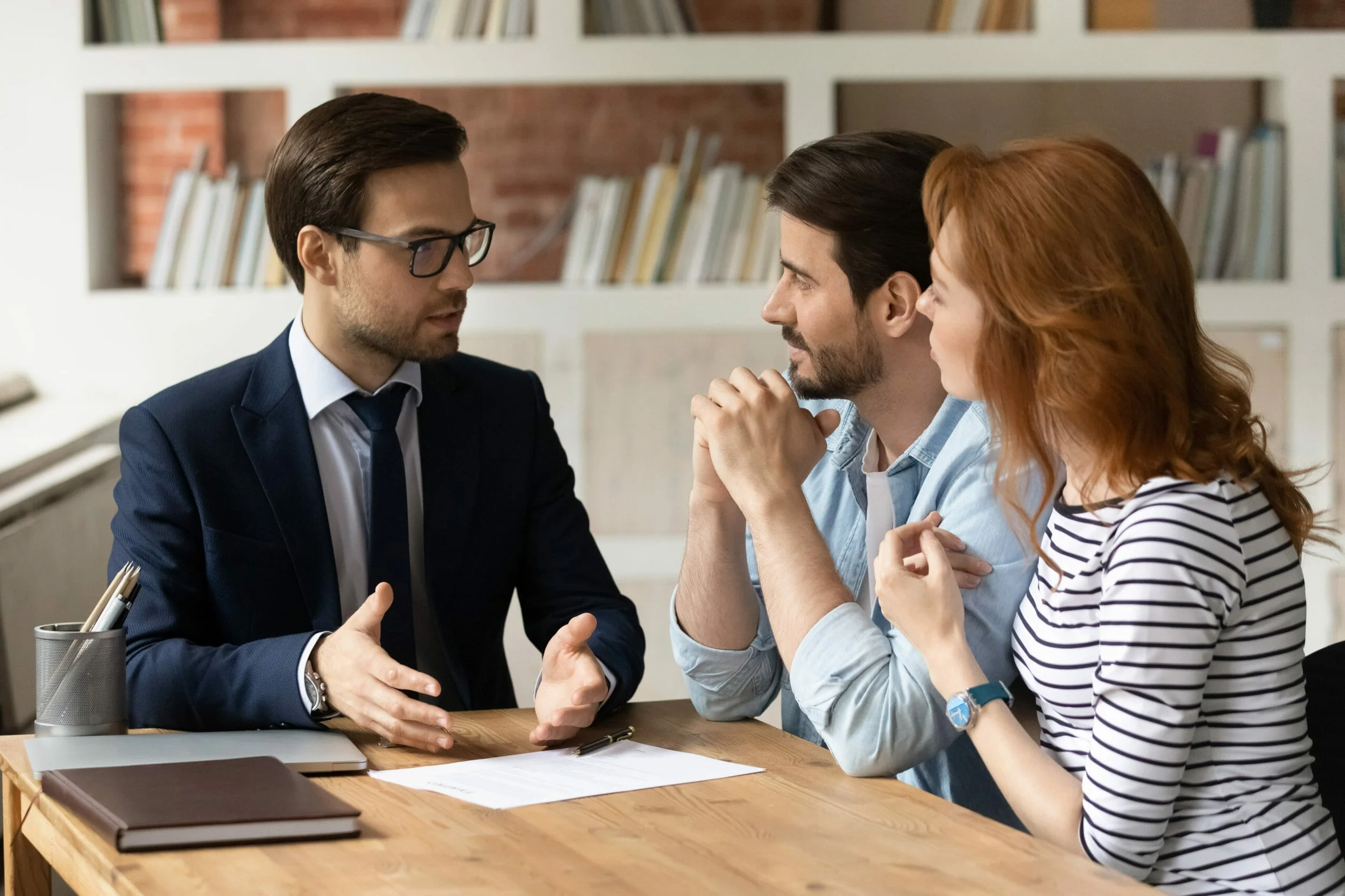 Business meeting with three people discussing at a wooden table in an office, with bookshelves and brick wall in background.