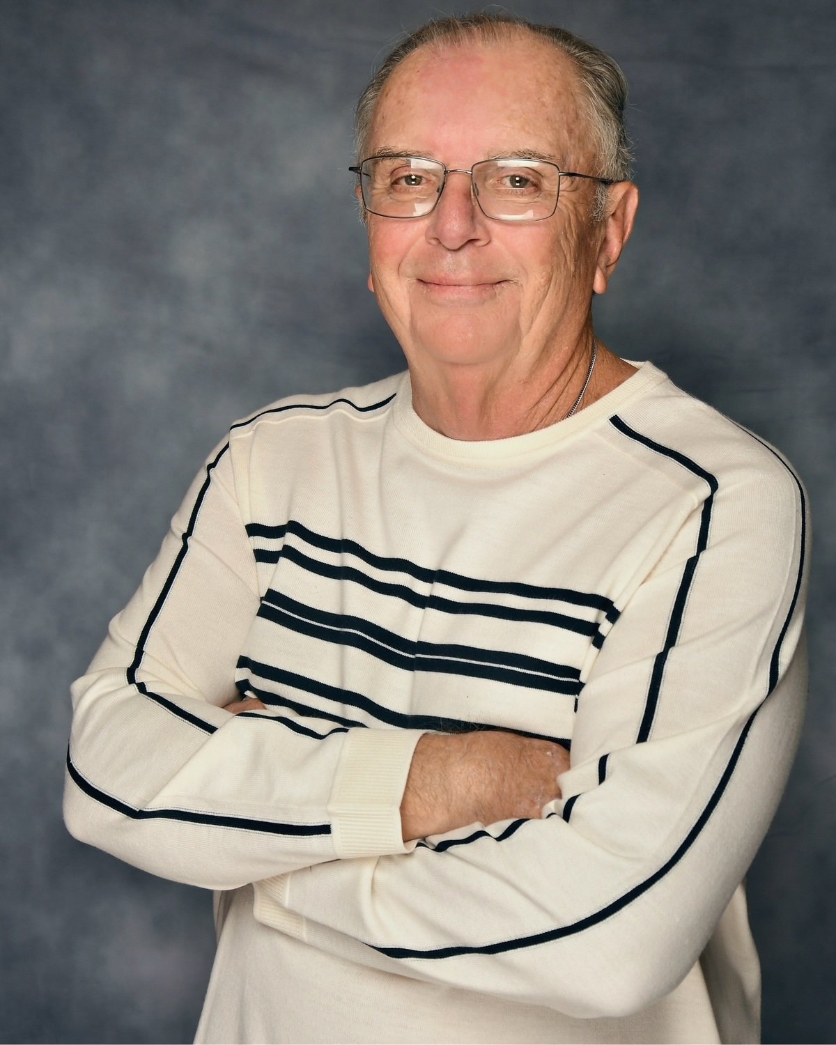 A smiling elderly man with glasses and gray hair, wearing a cream-colored sweater with black stripes, standing with his arms crossed against a gray background.
