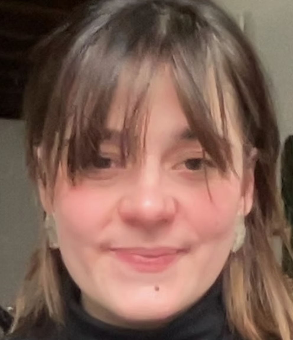 Smiling woman with shoulder-length brown hair and parted bangs, wearing a black turtleneck indoors.