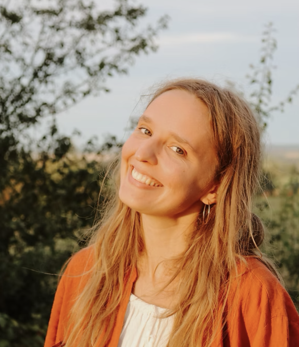 A woman with long, wavy red hair smiling outdoors, wearing a white top and an orange jacket with a background of trees and sky.