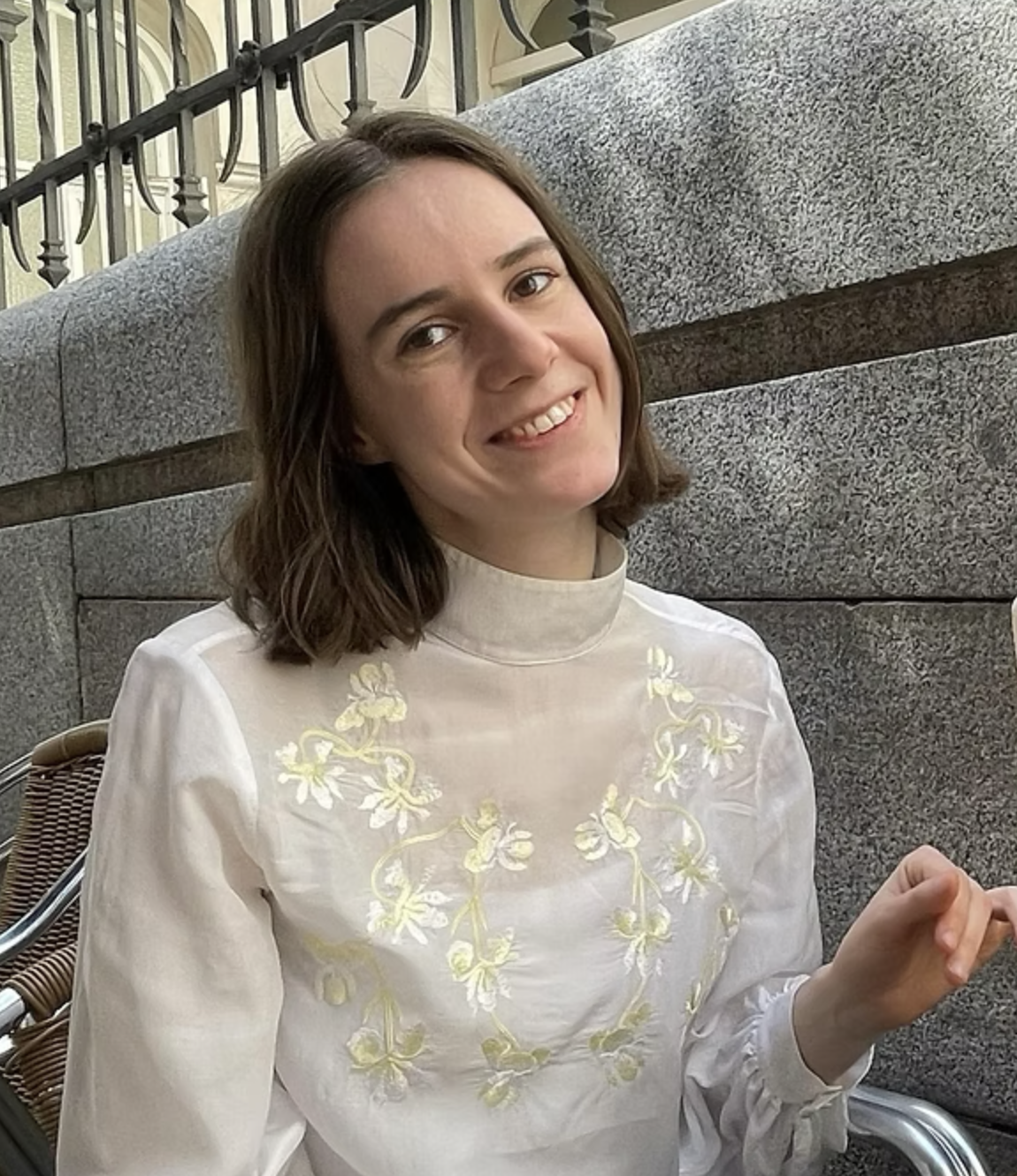 A woman with shoulder-length brown hair, smiling, wearing a light-colored embroidered blouse, sitting outdoors near a stone wall and a metal railing.