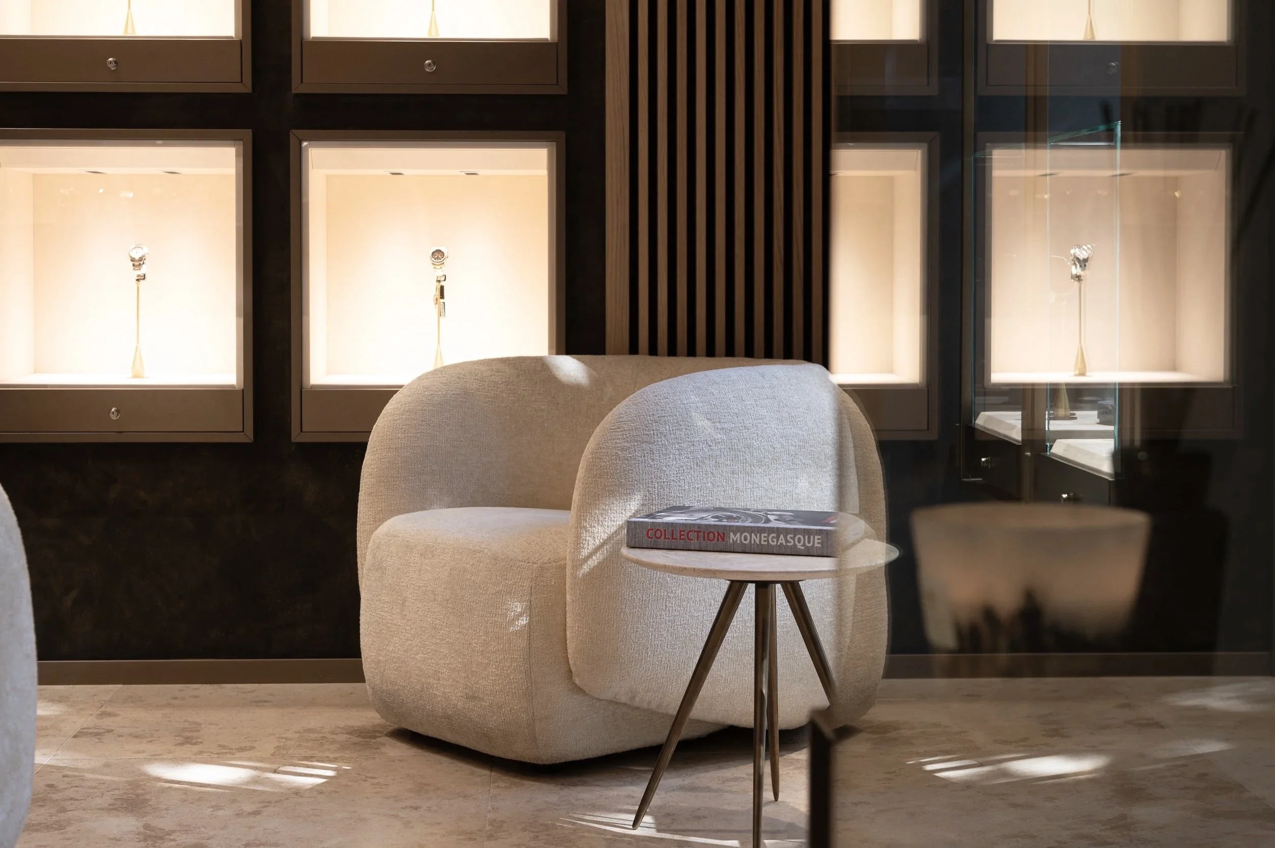 Costantini Watches Brand Identity by Define London - Real Estate A beige, rounded armchair next to a small round side table with a magazine on top, set in a modern decor with illuminated display cases in the background.