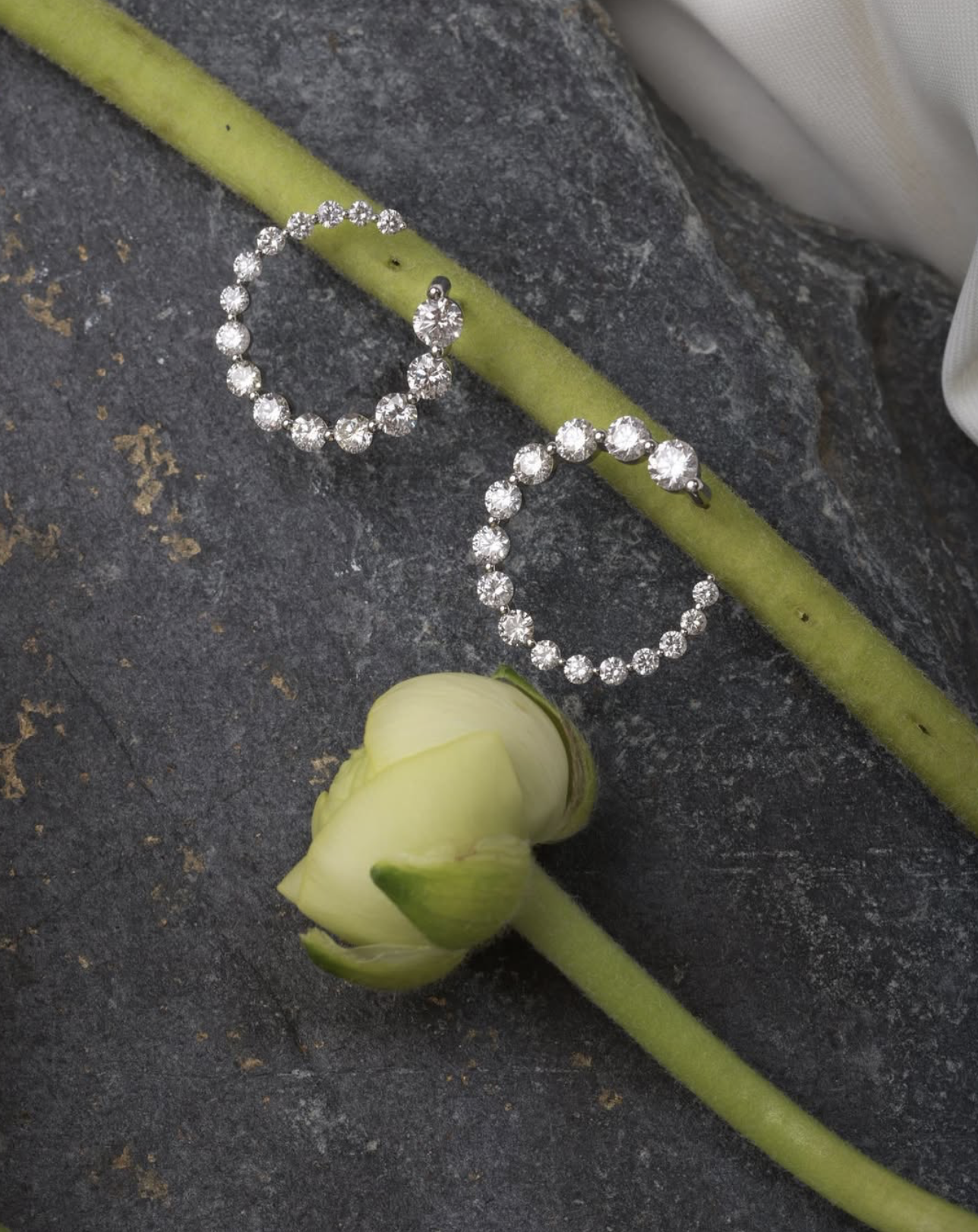 A pair of silver hoop earrings with clear gemstones, placed on a green stem of a white flower with a closed bud, on a dark textured surface.