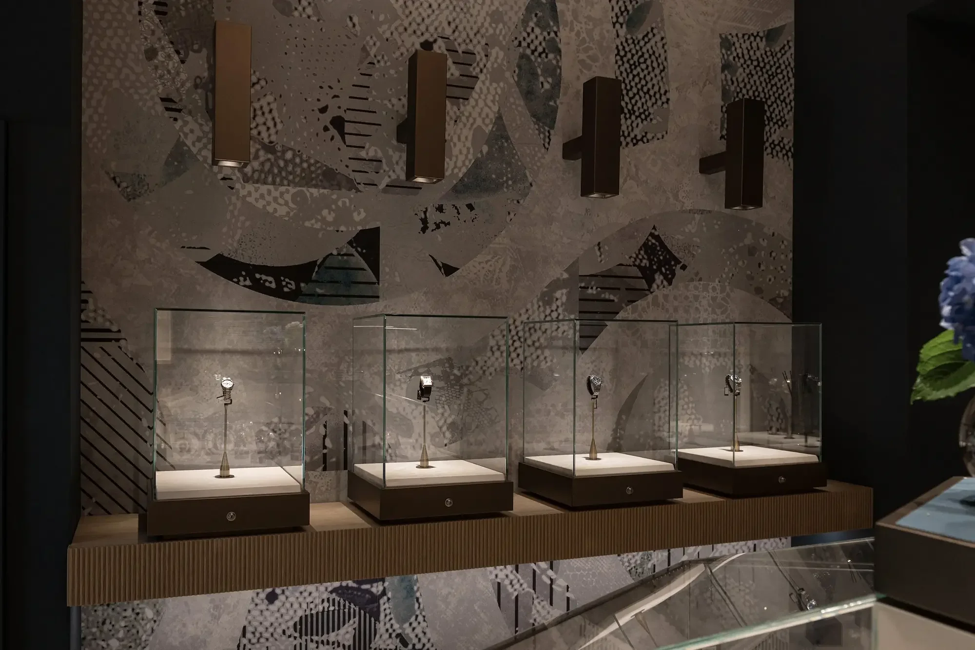 A display area in a retail store showcasing watches, with multiple watches mounted on individual stands behind glass. The background features a decorative abstract wall with geometric shapes and patterns.