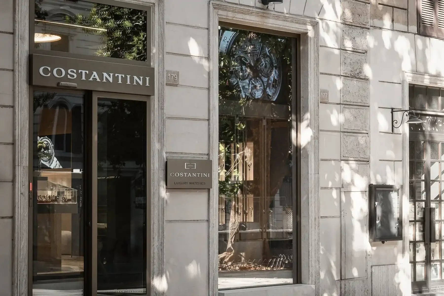 Storefront of Costantini luxury watch boutique with a glass window displaying watches, a sign reading 'COSTANTINI' above the door, and a small plaque indicating the store's name and specialization in luxury watches. The building has a stone facade wi