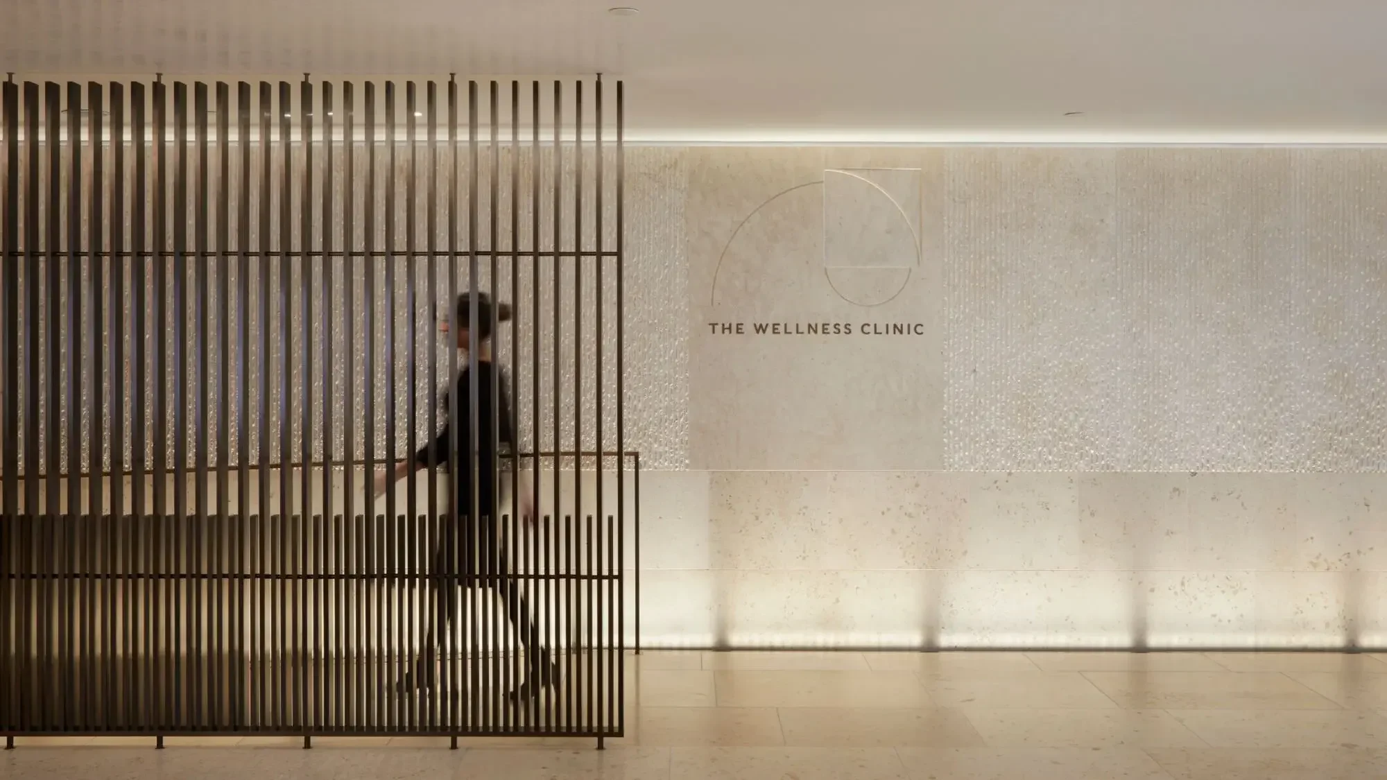 Person walking past a wooden slat partition at The Wellness Clinic, a modern interior with a wall featuring textured tiles and a logo of a circle with a square inside.