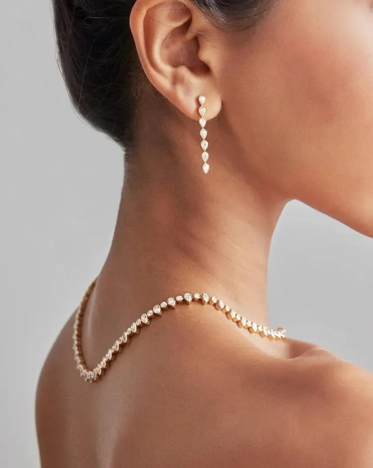 Close-up of a woman wearing diamond jewelry, including a long dangling earring and a pearl necklace, with her bare shoulder and neck visible.
