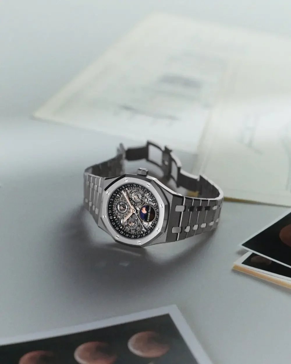 A silver wristwatch with a detailed black dial displaying multiple subdials and moon phase, placed on a white surface next to printed photographs and a blurred background of a computer or electronic device.
