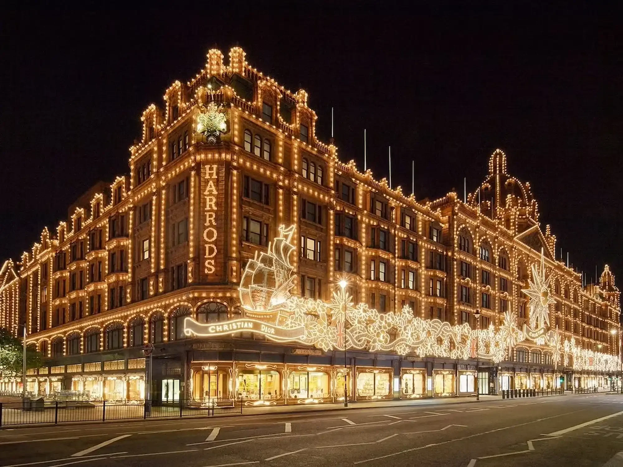 A multi-story building decorated with Christmas lights at night, with the words 'Harrods' and 'Christian Dior' illuminated. The building has an elaborate facade with illuminated outlines, snowflakes, and festive decorations, against a dark night sky.