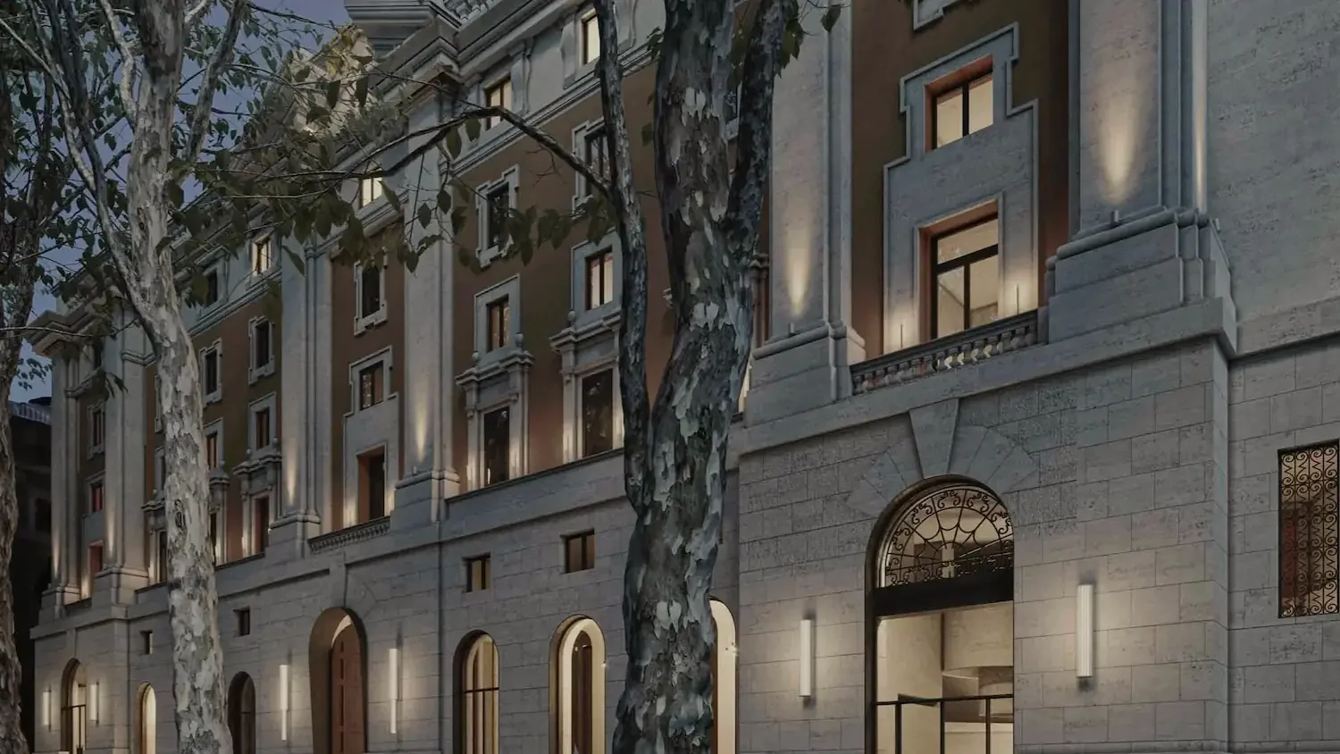 A historic building with large windows and ornate architectural details seen at dusk, with trees in front and lights illuminating the facade.