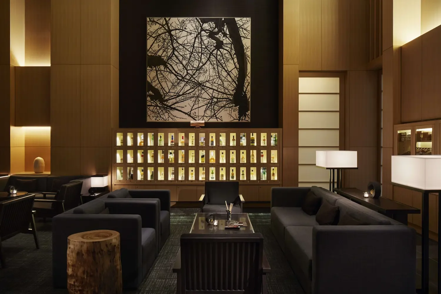 Interior of a modern, dimly lit lounge with gray sofas, black chairs, a wooden coffee table, and decorative lamps. A large artwork of tree branches is displayed on the wall above a backlit display of liquor bottles.