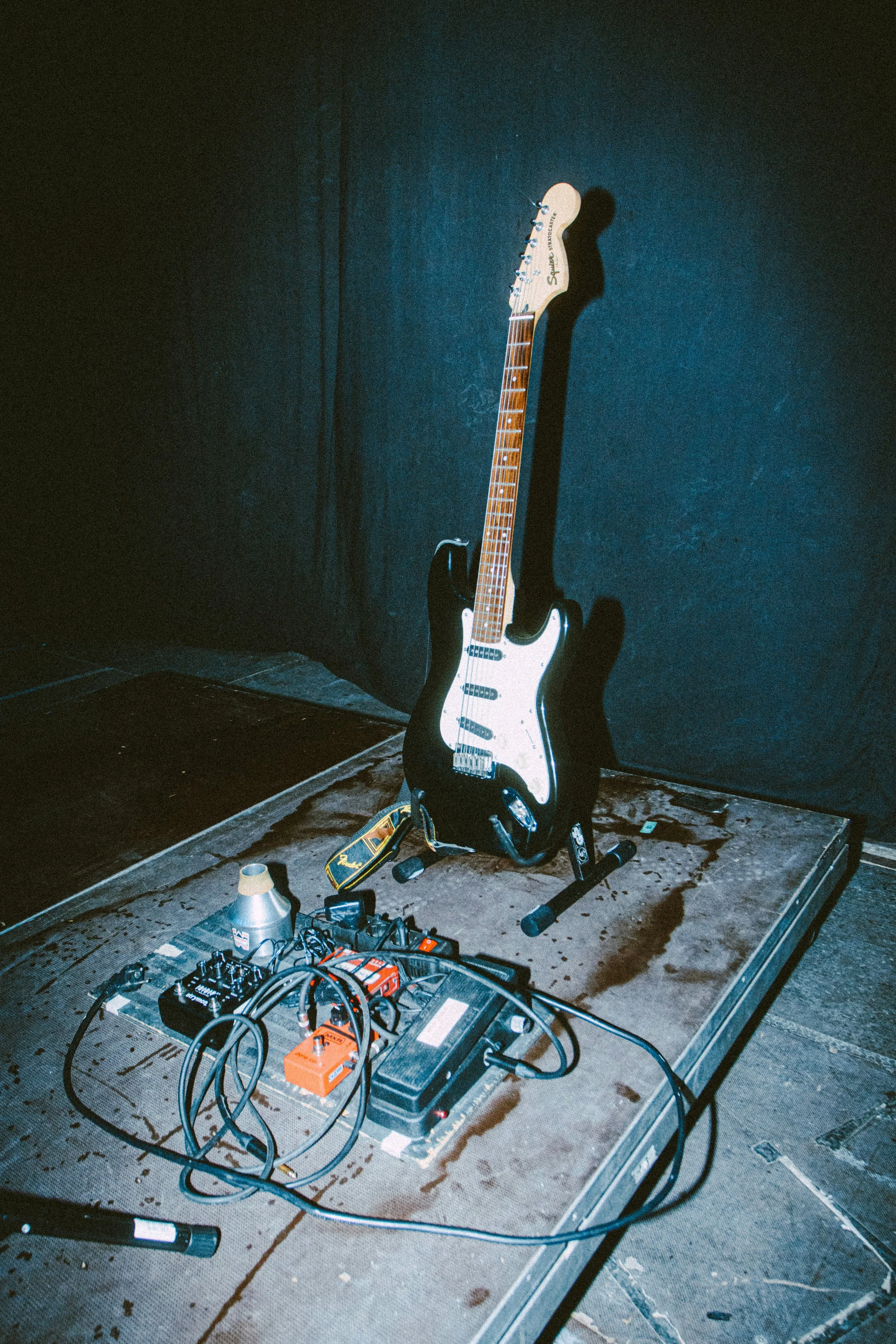 An electric guitar resting on a stand with effects pedals and equipment on the ground, against a dark backdrop.