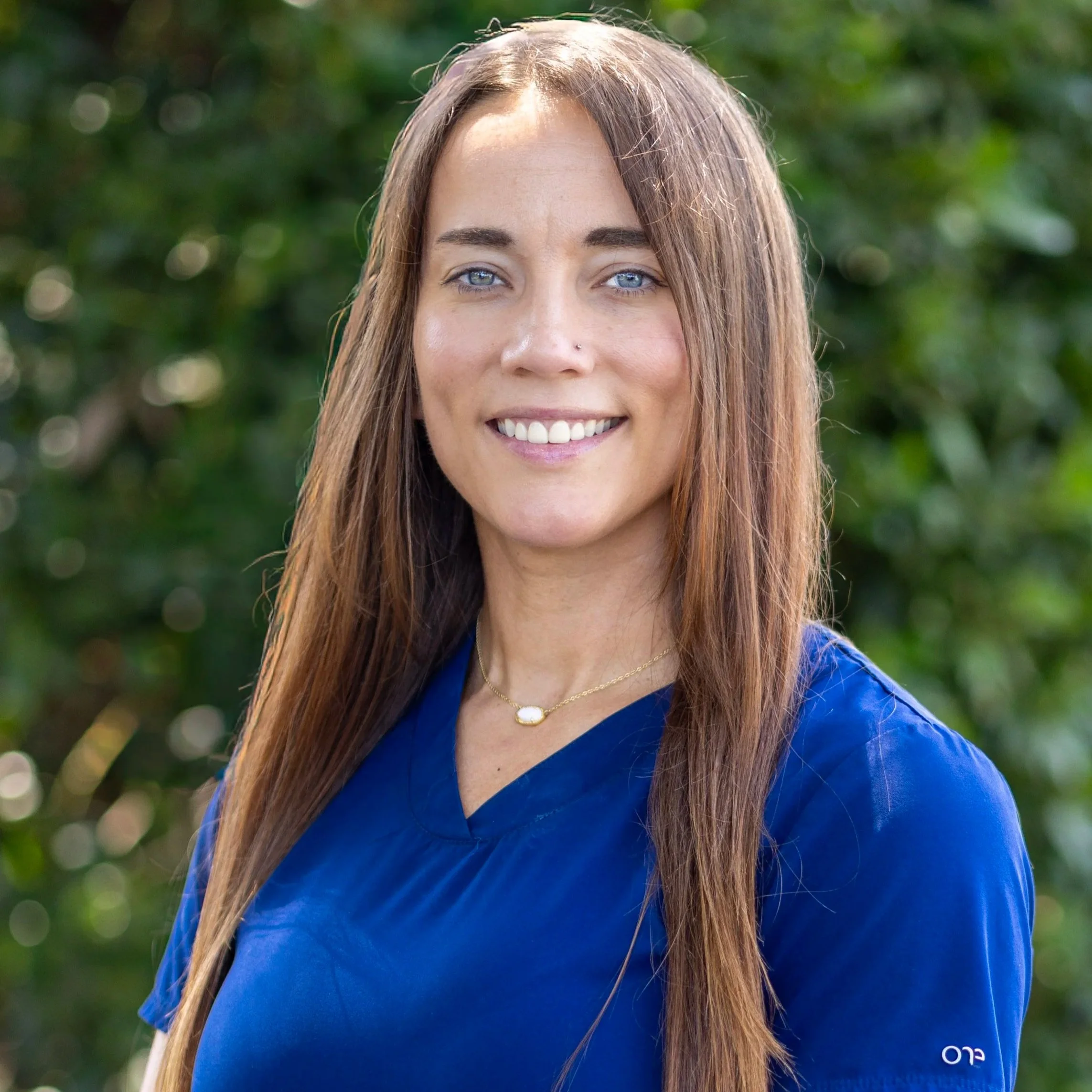 A woman with long, wavy, reddish-brown hair and blue eyes wearing a blue top and a necklace, smiling outdoors with greenery in the background.
