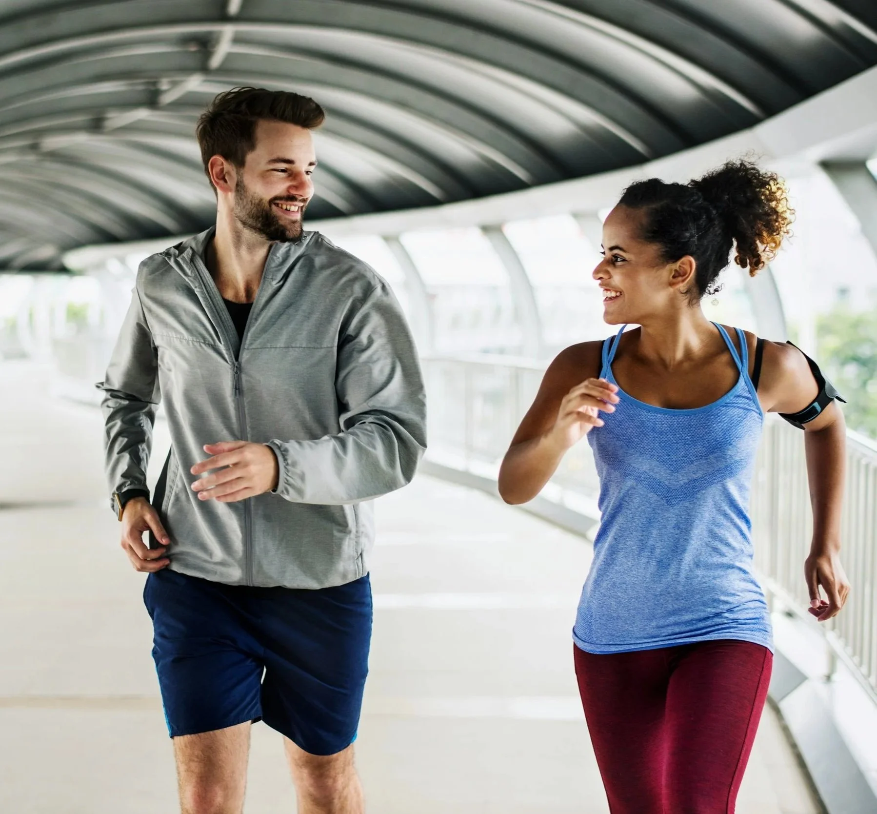A man and woman jogging together in a covered outdoor walkway, smiling and looking at each other.
