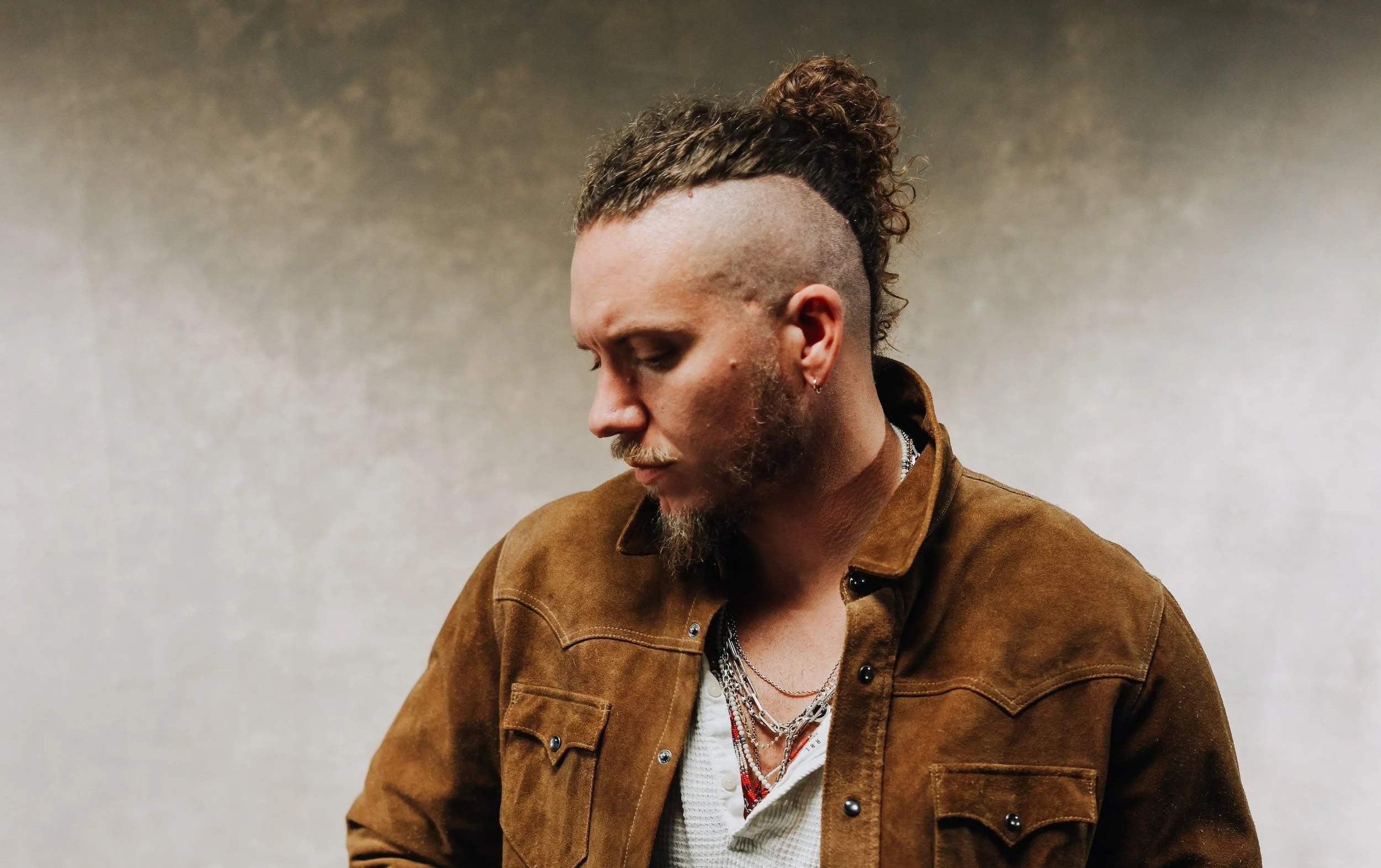 A man with a unique hairstyle, including a mohawk with dreadlocks on top, a shaved side, a beard, and multiple necklaces, looks downward against a plain gray wall.