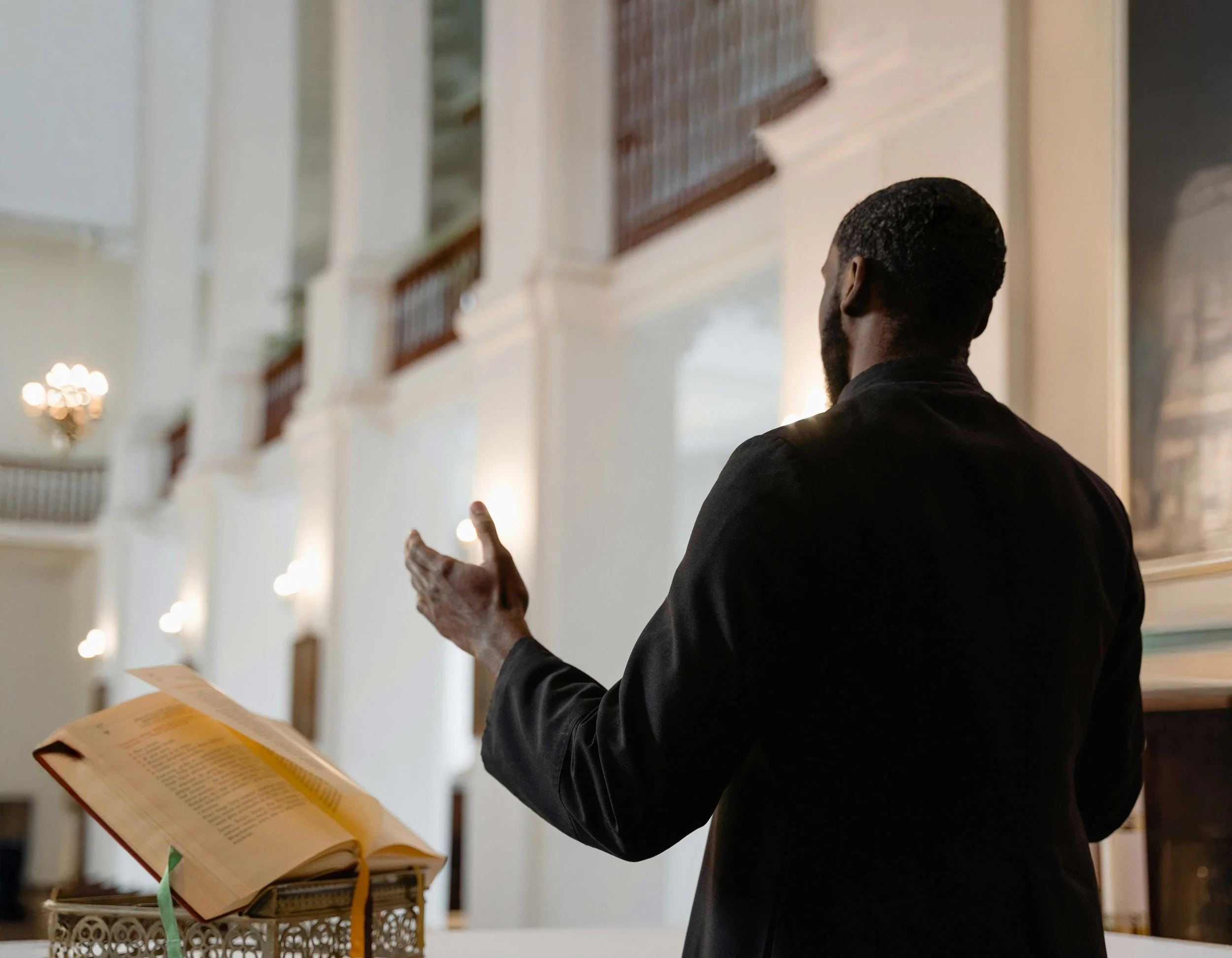 A man with dark hair and beard, wearing a black jacket, is standing in a church or chapel, facing away, with his right hand raised. An open book is on a decorative stand in front of him, with a green ribbon bookmark.