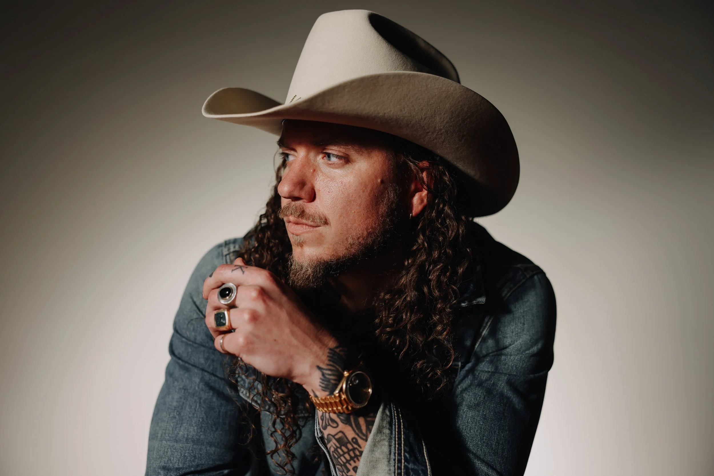 A man with a unique hairstyle, including a mohawk with dreadlocks on top, a shaved side, a beard, and multiple necklaces, looks downward against a plain gray wall.
