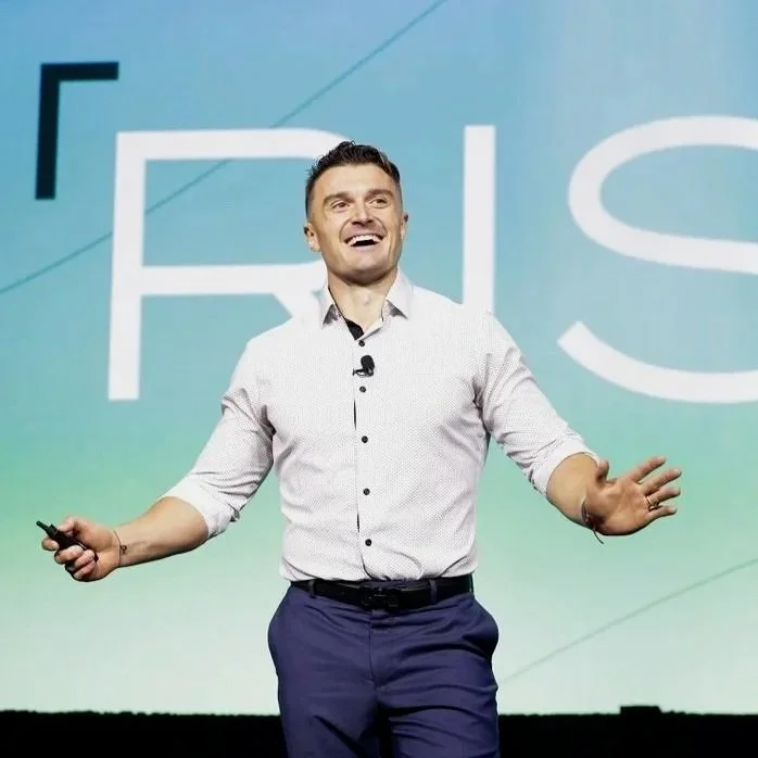 A man in a white shirt and dark pants is standing on stage, smiling, with one hand open and the other holding a remote or clicker. The background features a large screen with the partially visible word 'FIRS' in white letters on a light blue-green gradient.